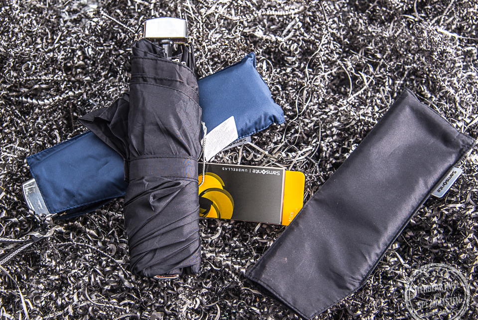 Schwarzer Mini Taschenschirm liegt auf dem blauen Pendant dazu. Die H&uuml;lle des schwarzen Schirms liegt daneben.