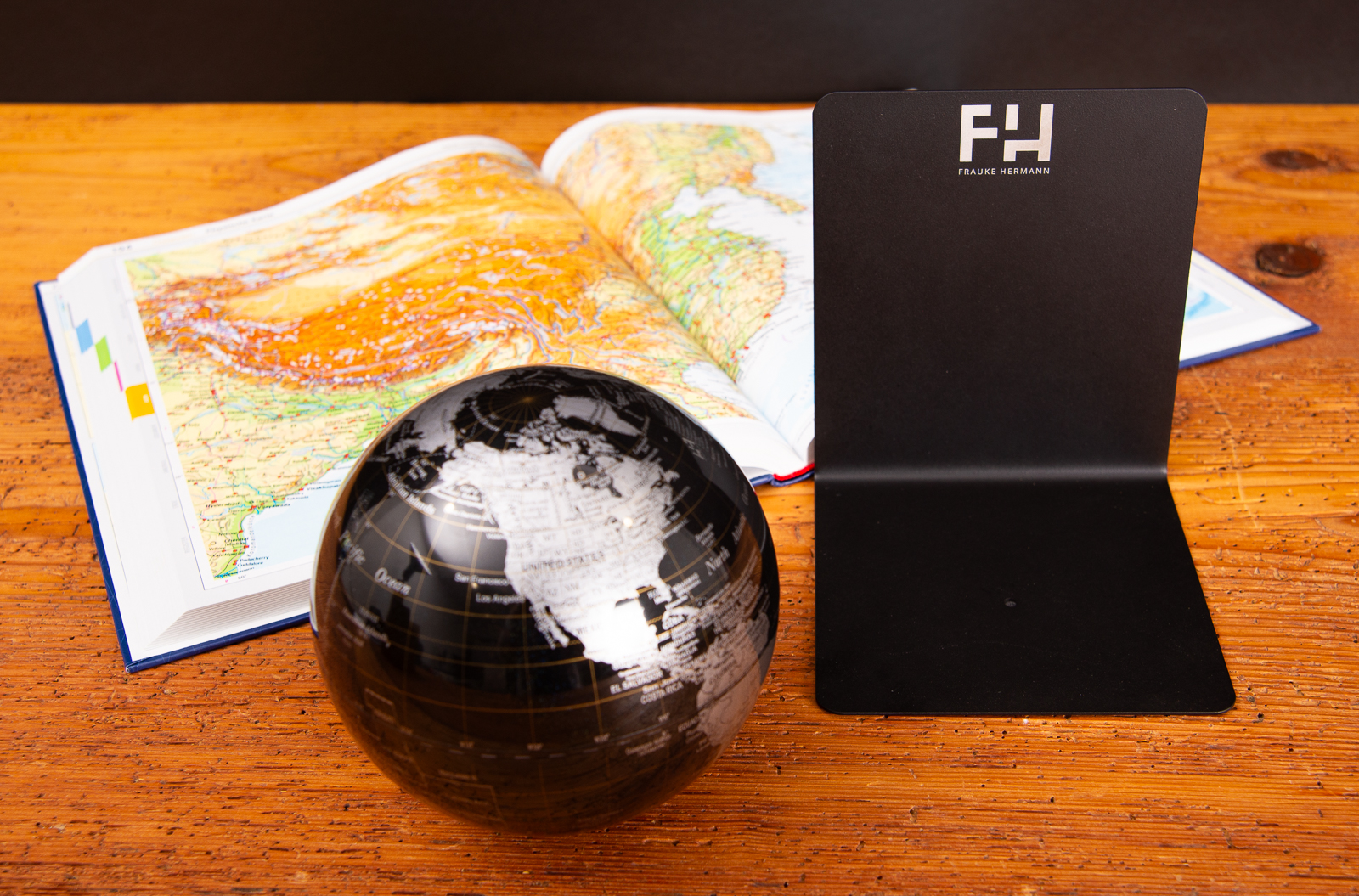Troika Globus und mattschwarze Buchst&uuml;tze mit Logogravur neben aufgeschlagenem Diercke Atlas auf Holztisch.