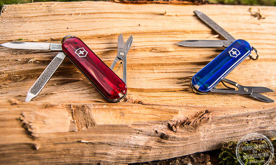 Zwei Victorinox Taschenmesser Classic SD aufgeklappt auf Holz liegend als Werbeartikel.