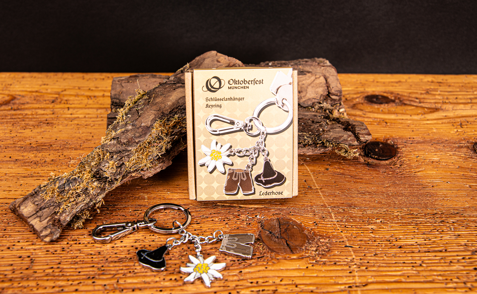 Troika Schl&uuml;sselanh&auml;nger mit Lederhose, Enzian und Seppelhut auf Holztisch inklusive Verpackung als Werbeartikel.