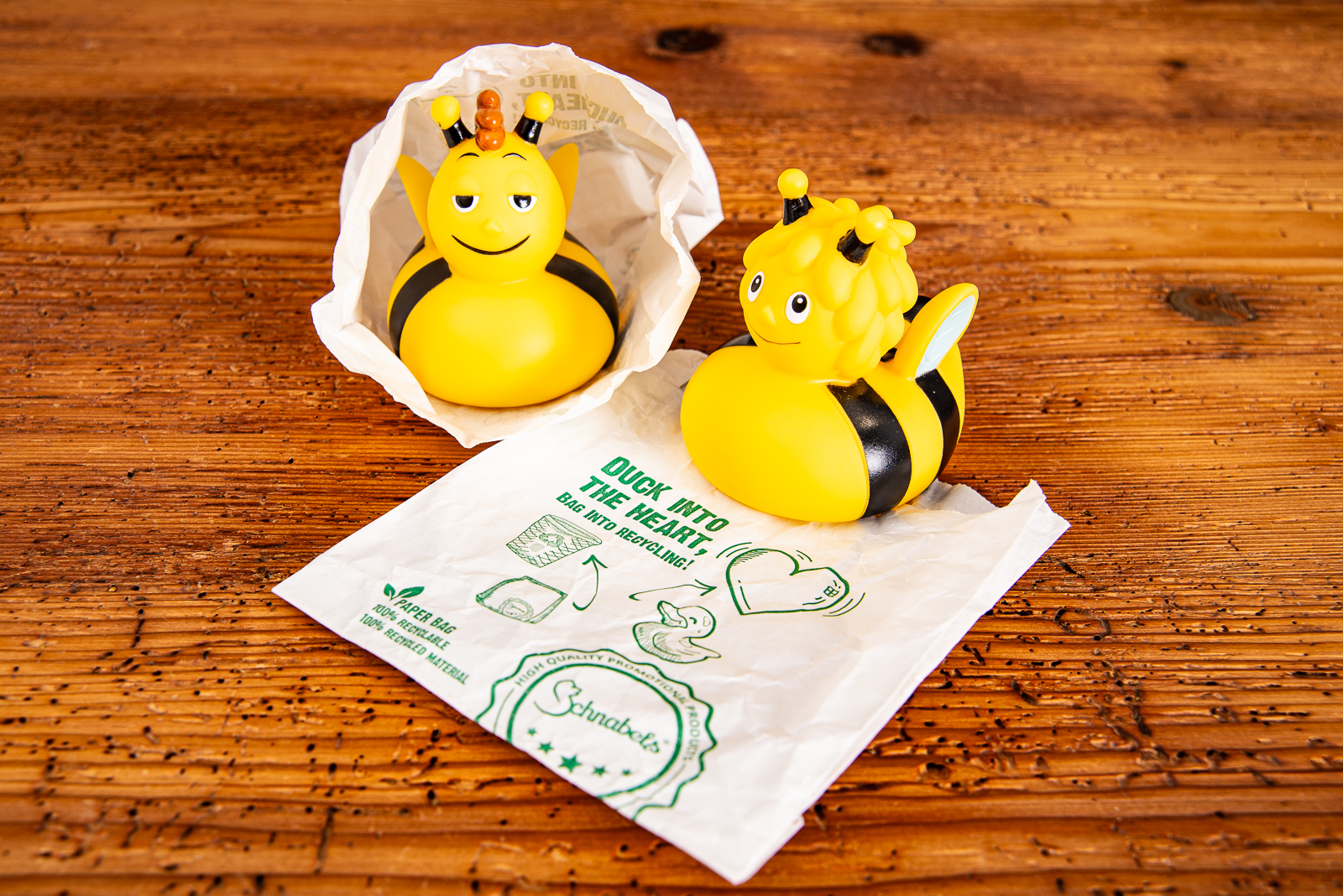 Quietscheenten Biene Maja und Biene Willi in Verpackungst&uuml;te auf Holztisch.