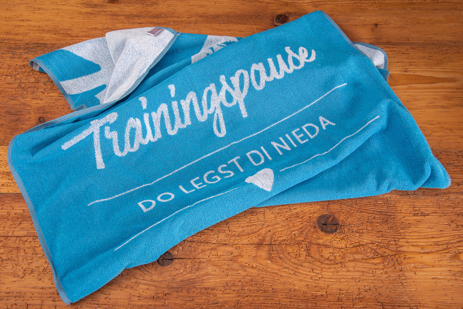 Badetuch mit Logoeinwebung als Werbeartikel im Sport und Freizeitbereich auf Holzboden.