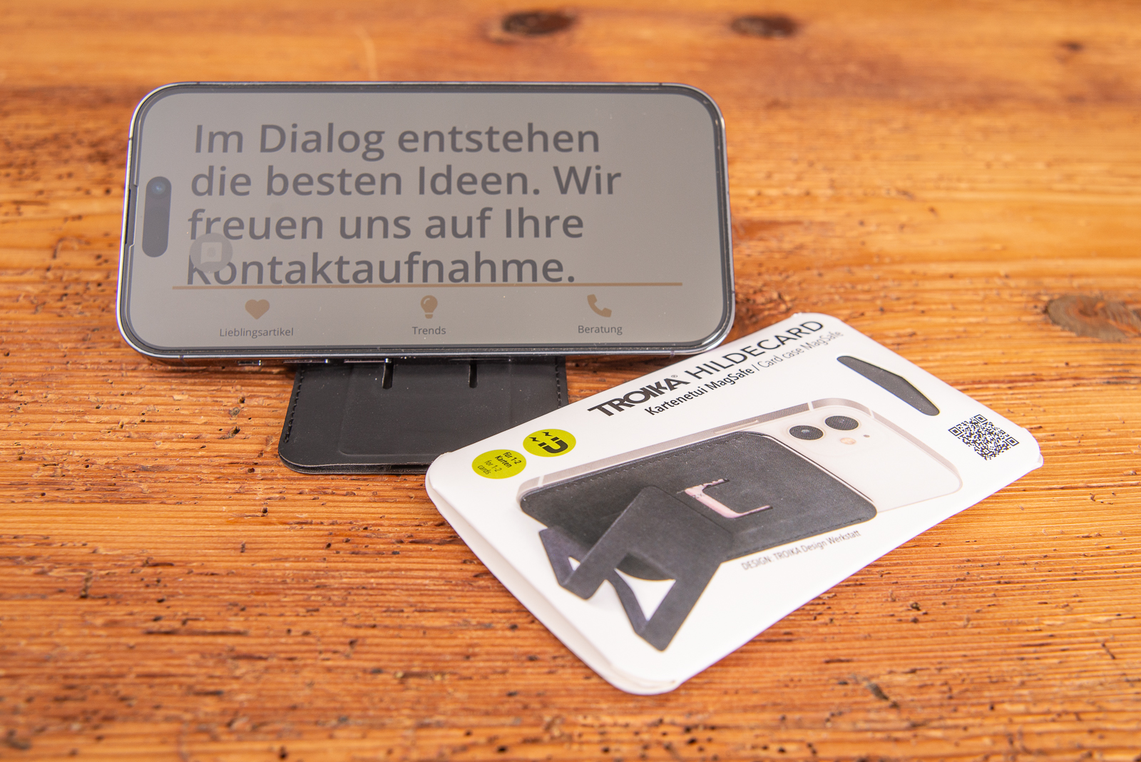 Das Troika Hildecard ist Handyhalter und Kartenetui in einem und somit ein hochwertiger Werbeartikel aus Kunstleder f&uuml;r Mitarbeiter und Gesch&auml;ftskunden mit Verpackung auf Holztisch. 