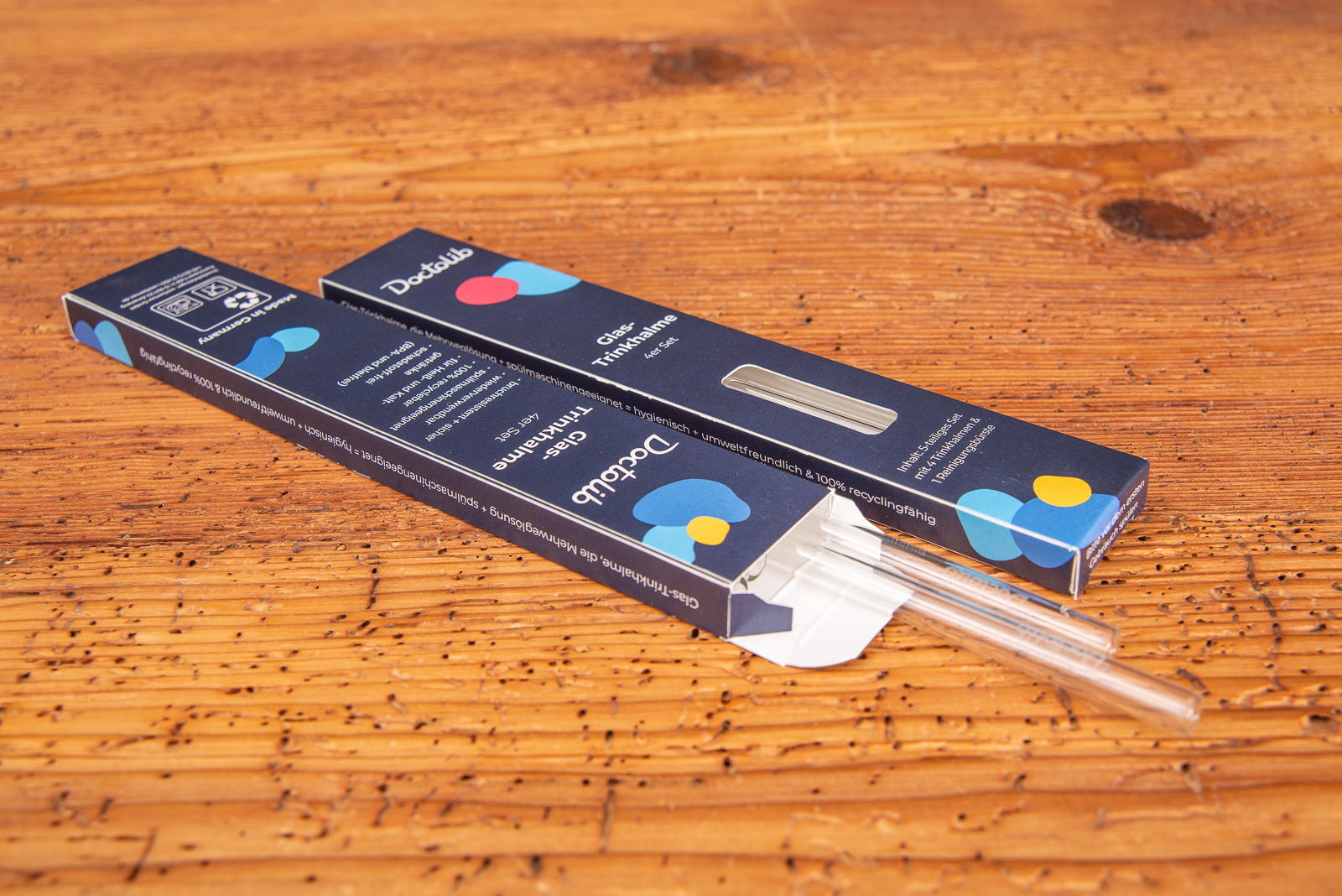 Zwei individuell bedruckte Glastrinkhalme mit Gravur als Werbegeschenk auf Holztischplatte.