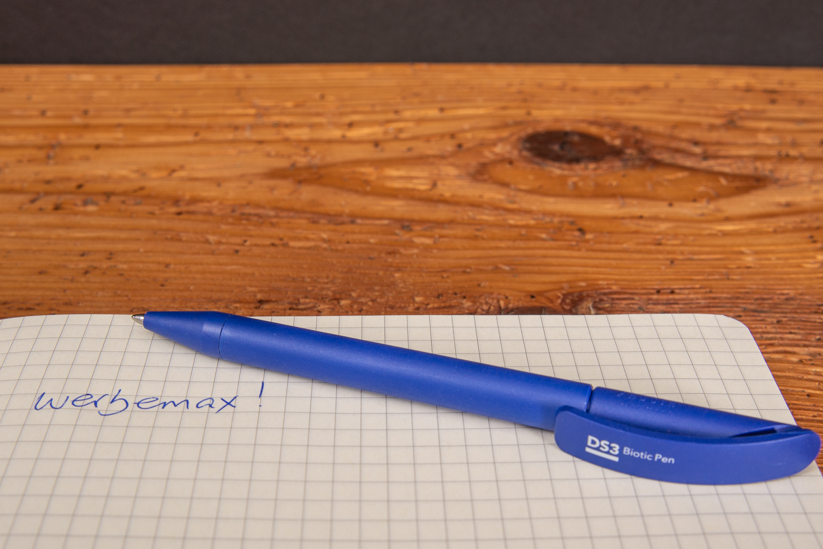 Ein blauer Werbekugelschreiber mit Logo liegend auf einem karierten Blatt Papier.