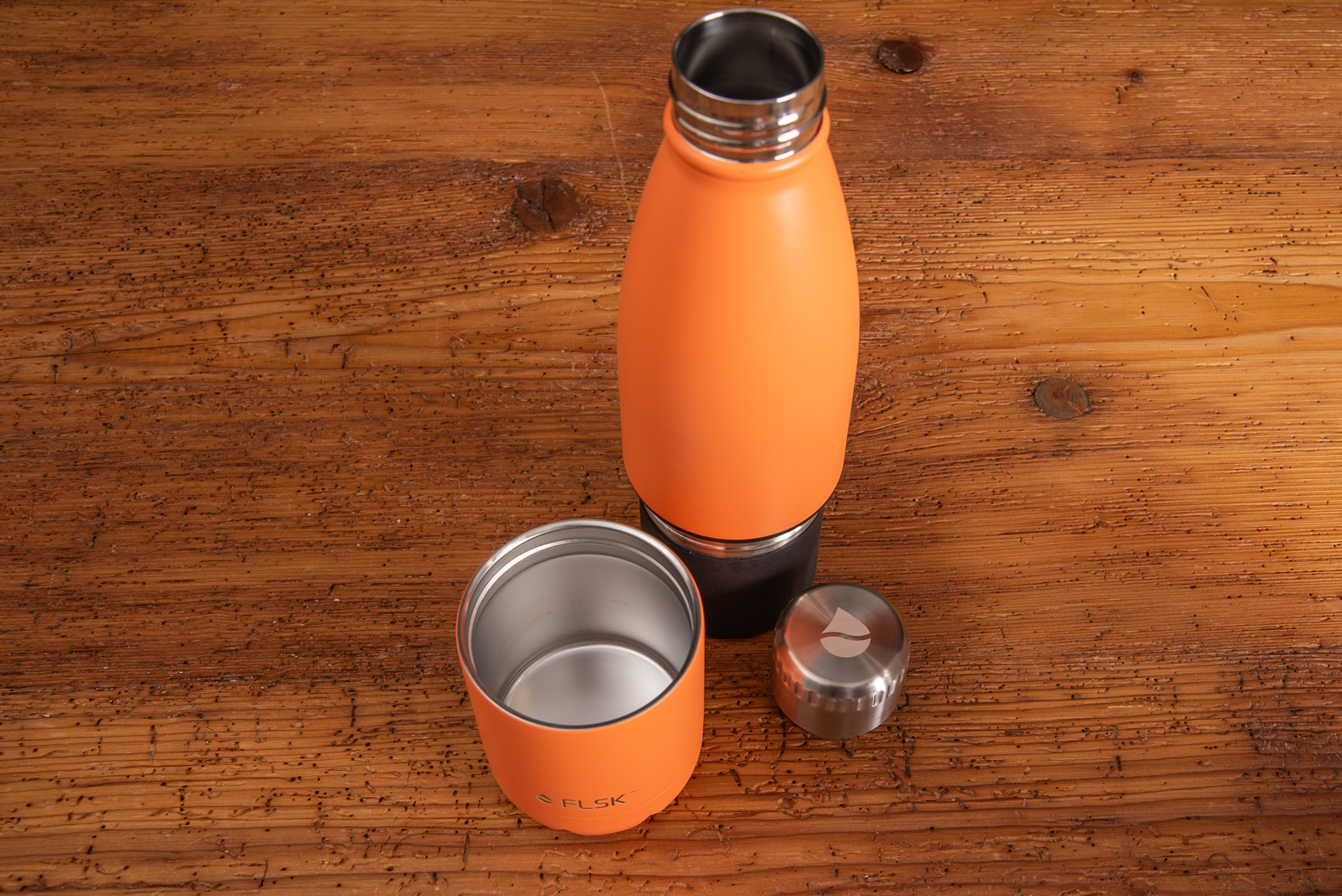 Trinkflasche mit Becher in Orange von dem Markenhersteller FLSK mit Logogravur auf Holztisch