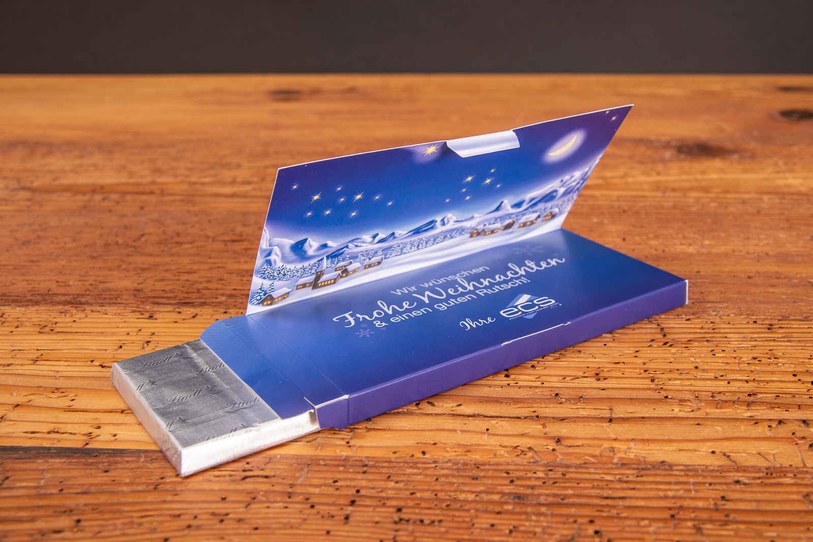 Individuell bedruckte Schokogru&szlig;-Klappkarte als Werbegeschenk Referenz ecs auf Holztisch.