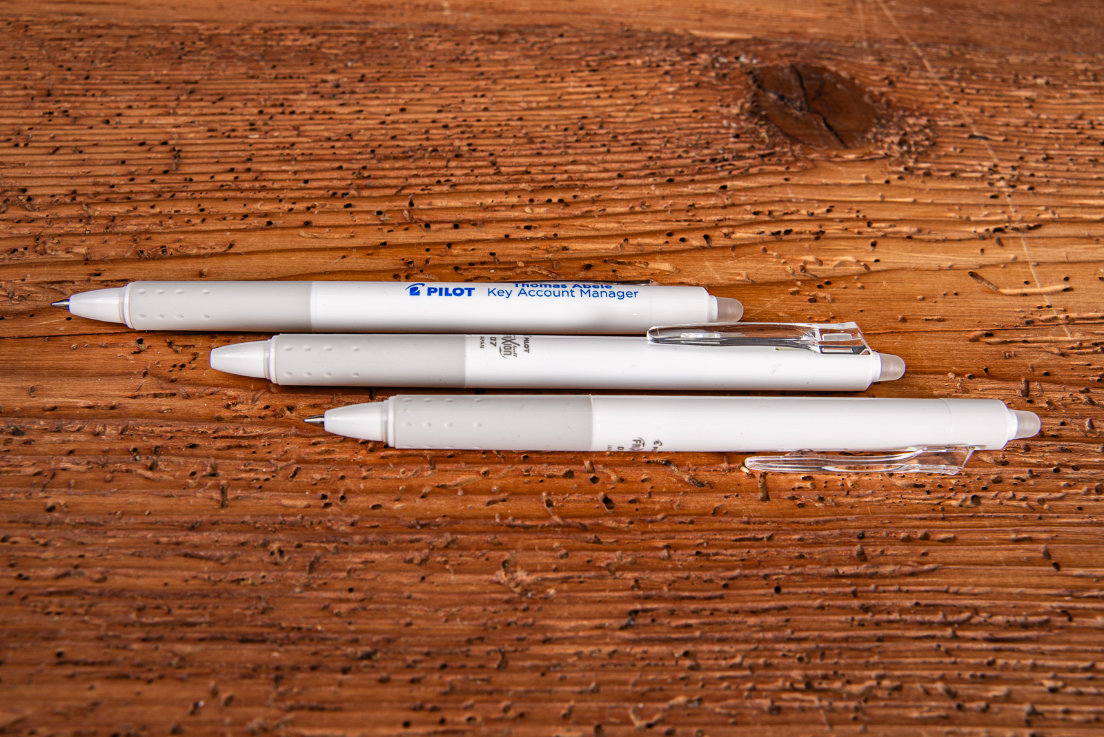 3 wei&szlig;er Frixion Clicker no Print mit einer gummierten Griffzone und Logodruck als Werbemittel im Merchandise auf Holztisch liegend. 