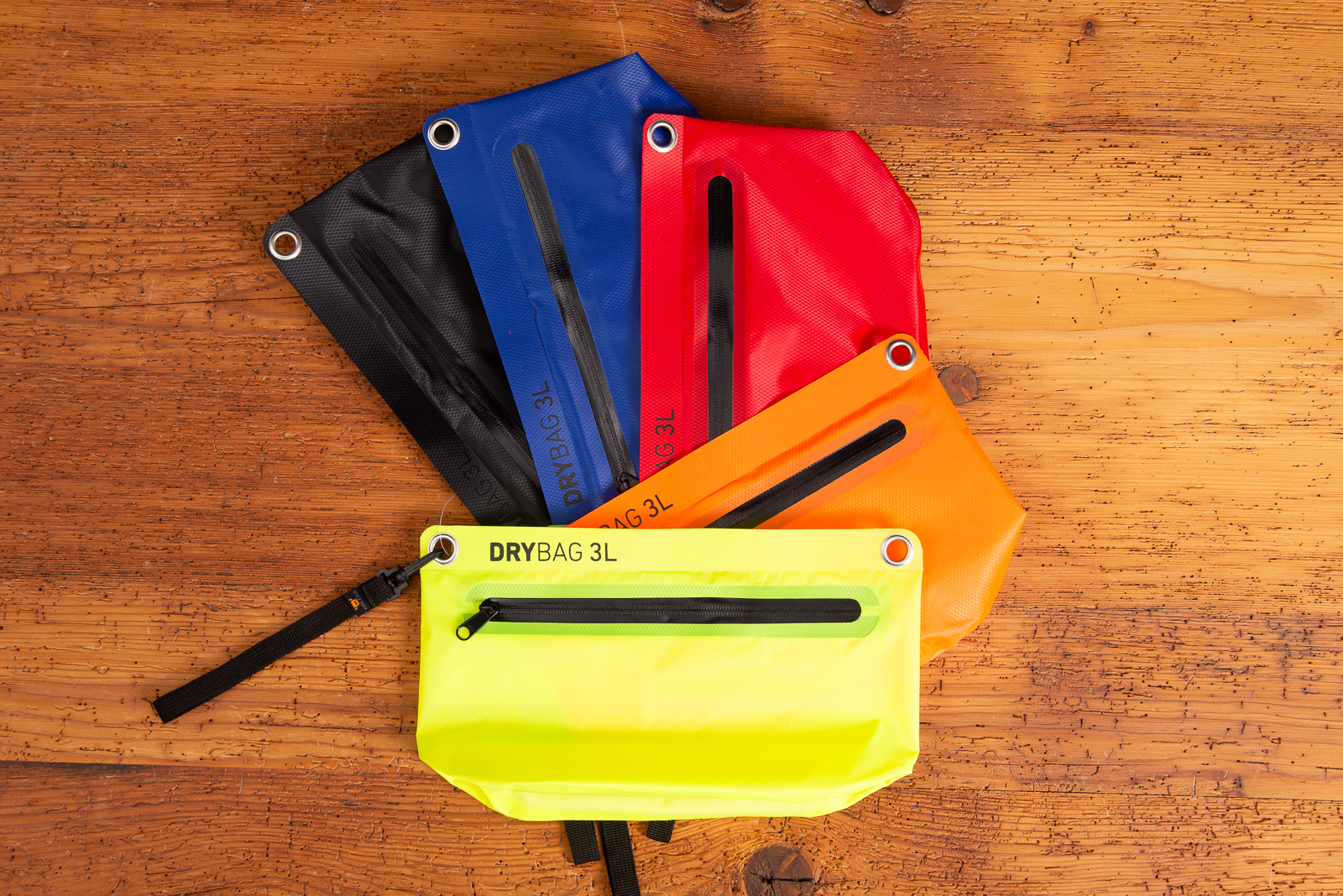 Farb&uuml;bersicht der Halfar Drybag 3L in Schwarz, Blau, Rot, Orange und Neongelb mit H&auml;ngeschlaufe aufgef&auml;chert auf Holzplatte liegend