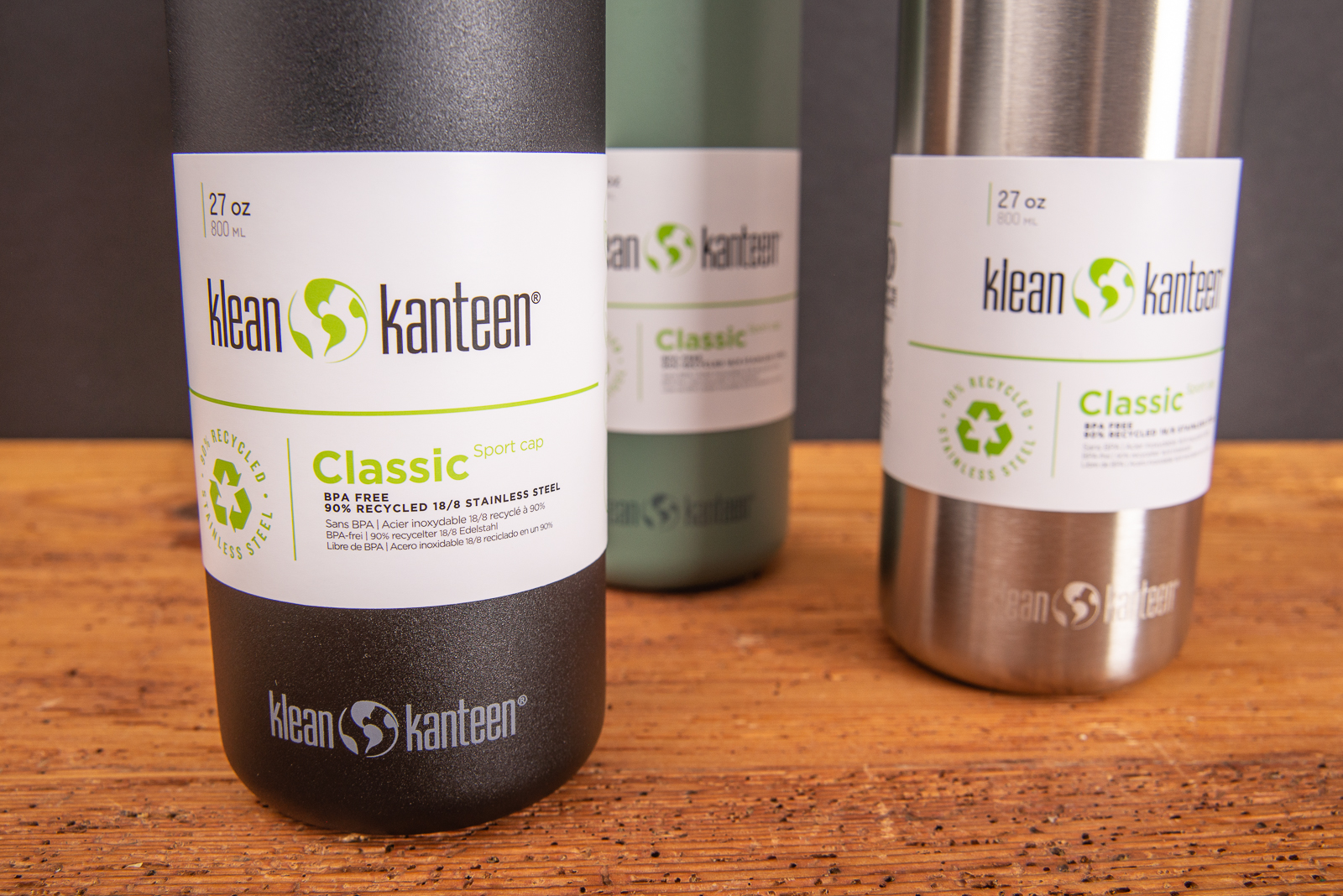 Klean Kanteen Edelstahlflaschen in schwarz, khaki und silber mit Logo graviert als nachhaltiges Merch f&uuml;r Kunden und Mitarbeiter auf Holztisch