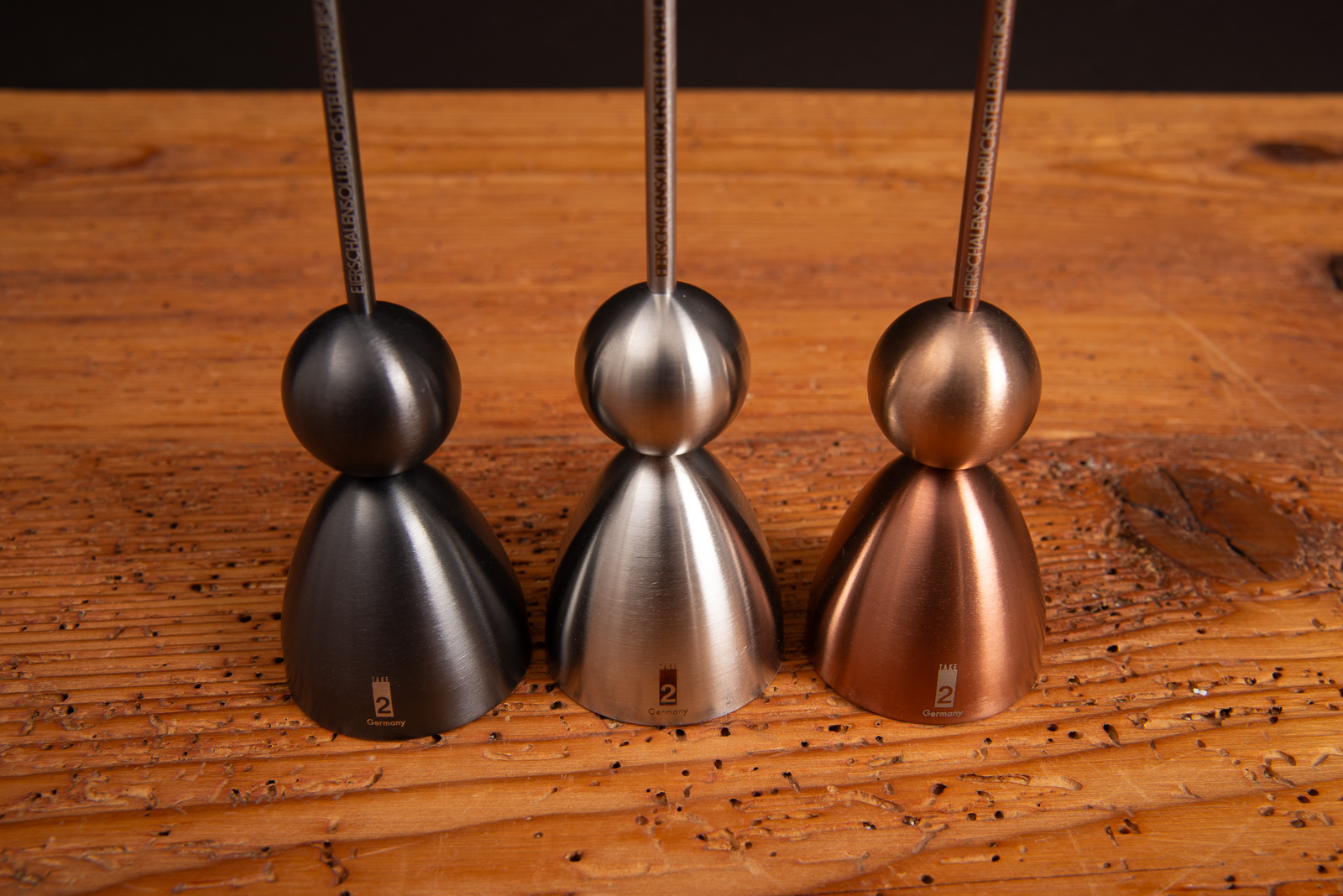 Clack Eierk&ouml;pfer von take2 im Farbvergleich &ndash; Schwarz, Silber und Kupfer auf Holzoberfl&auml;che.