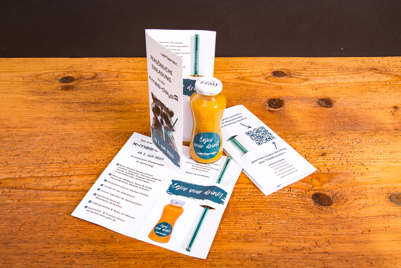Einladung zu den werbemax xmasdays 2025 mit Flyer, Glasstrohhalm, Smoothieflasche mit &bdquo;Enjoy your drink&ldquo;-Aufdruck und QR-Code &ndash; kreative Werbeartikel-Pr&auml;sentation auf Holztisch.
