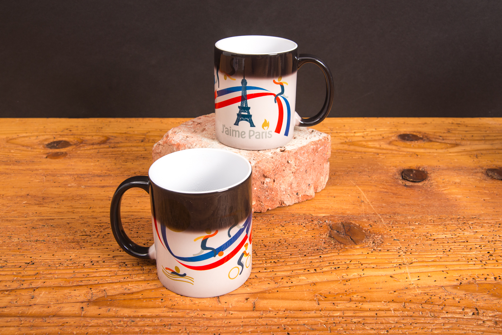 Zwei Magic-Tassen mit Farbwechsel und individuellem Aufdruck &ndash; Werbeartikel Tassen mit Thermoeffekt von werbemax auf Holztisch und Backstein platziert.