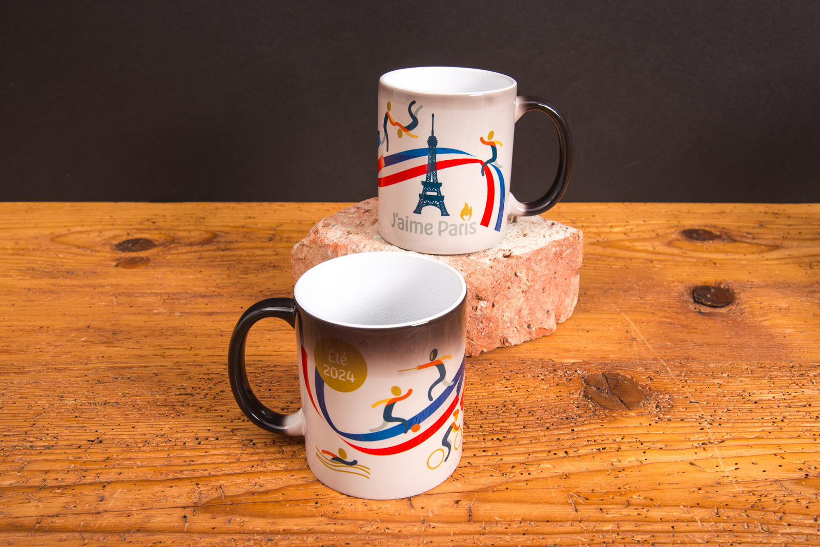 Zwei Magic-Tassen mit Farbwechsel-Design &bdquo;J&rsquo;aime Paris&ldquo; zeigen das Motiv durch hei&szlig;e Fl&uuml;ssigkeit &ndash; bedruckte Werbeartikel Tassen mit Thermoeffekt von werbemax auf Backstein.