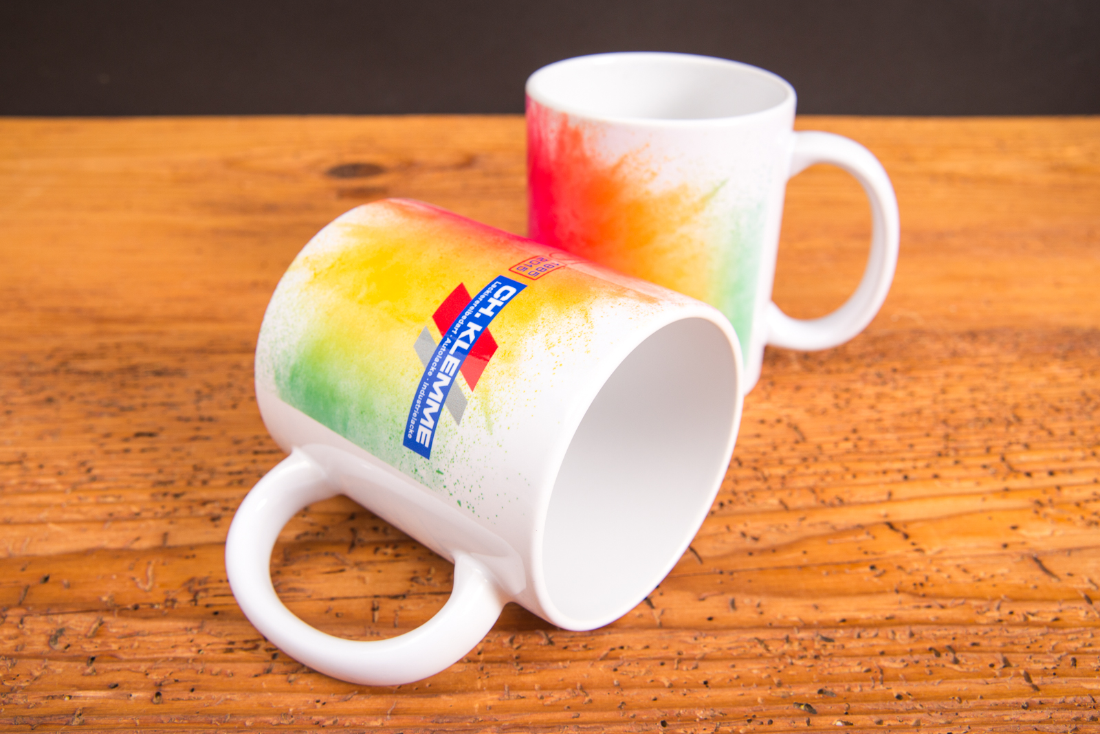 Zwei wei&szlig;e Tassen mit farbenfrohem Sublimationsdruck und Firmenlogo &ndash; individuell gestaltete Werbeartikel Tassen mit Regenbogendesign.