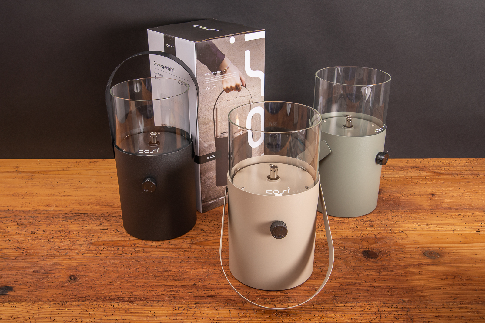 Cosiscoop Original Gaslaternen in Schwarz, Beige und Olivgr&uuml;n mit Verpackung auf Holztisch, ein hochwertiges Outdoor-Licht als Werbegeschenk mit Logo.