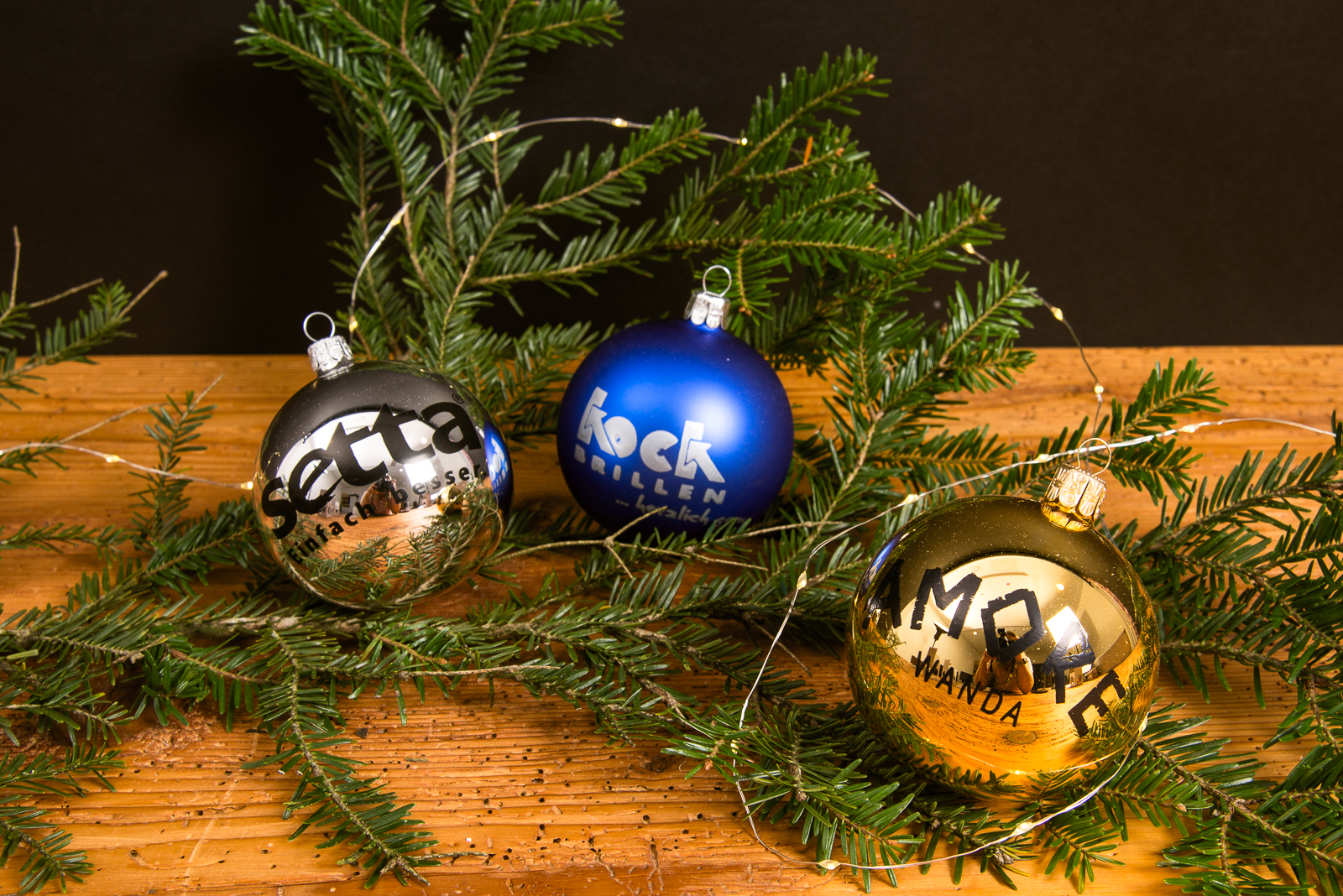 Personalisierte Weihnachtskugeln in Silber, Blau und Gold mit Logo, dekoriert an Tannenzweigen auf Holzplatte &ndash; individuelle Christbaumkugeln als Kundengeschenk zu Weihnachten.
