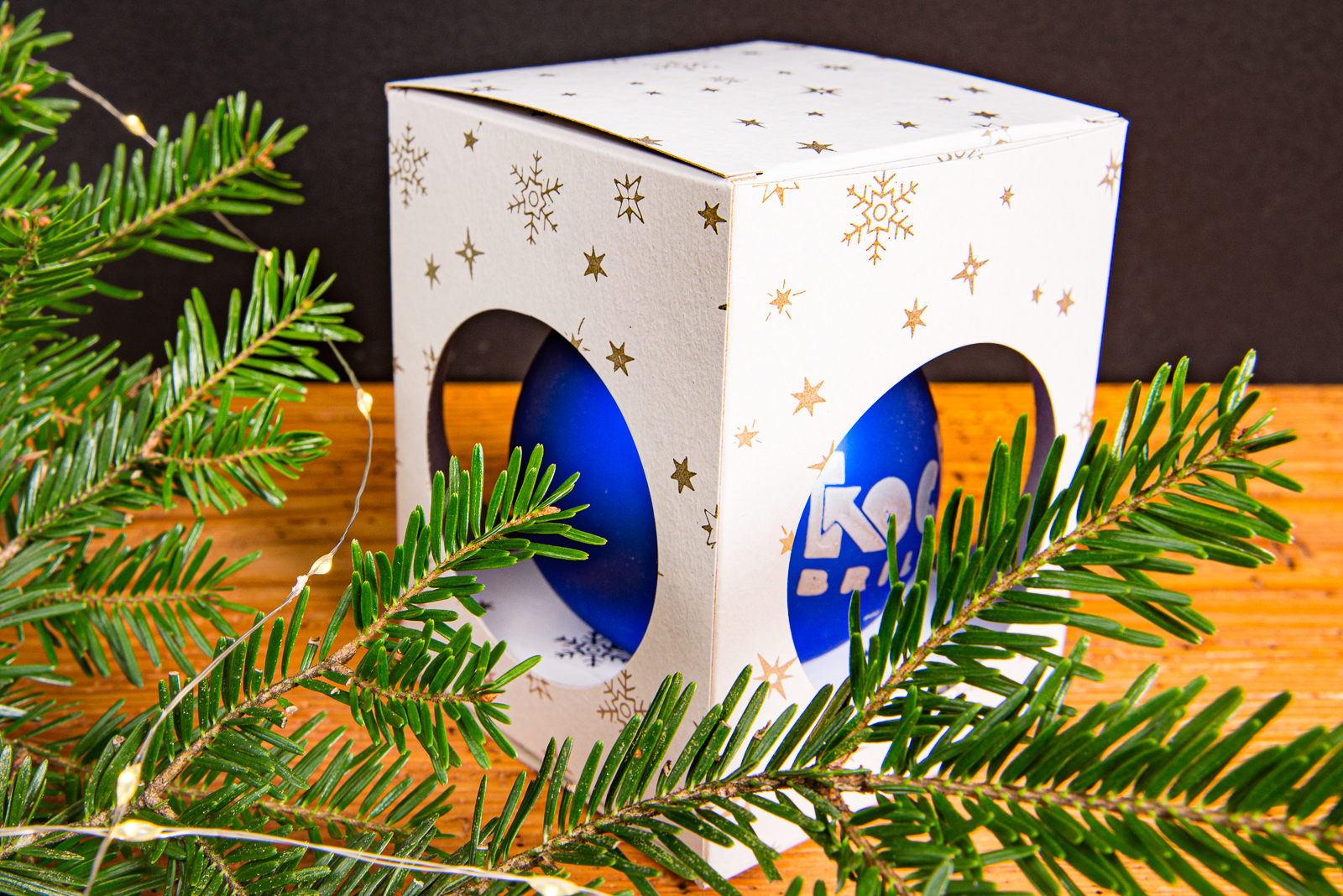 Blaue Weihnachtskugel mit Logo in wei&szlig;er Geschenkbox mit goldenen Sternen dekoriert mit Tannenzweigen als Merchandise Geschenk f&uuml;r Sport- und Musik-Fans.