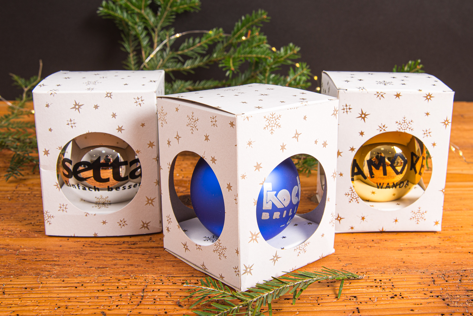 Silberne, blaue und goldene Weihnachtskugeln mit Logo in wei&szlig;er inzelverpackung mit Sternen als Werbegeschenke und elegante Kundengeschenke mit Tannenzweig auf Holzplatte.