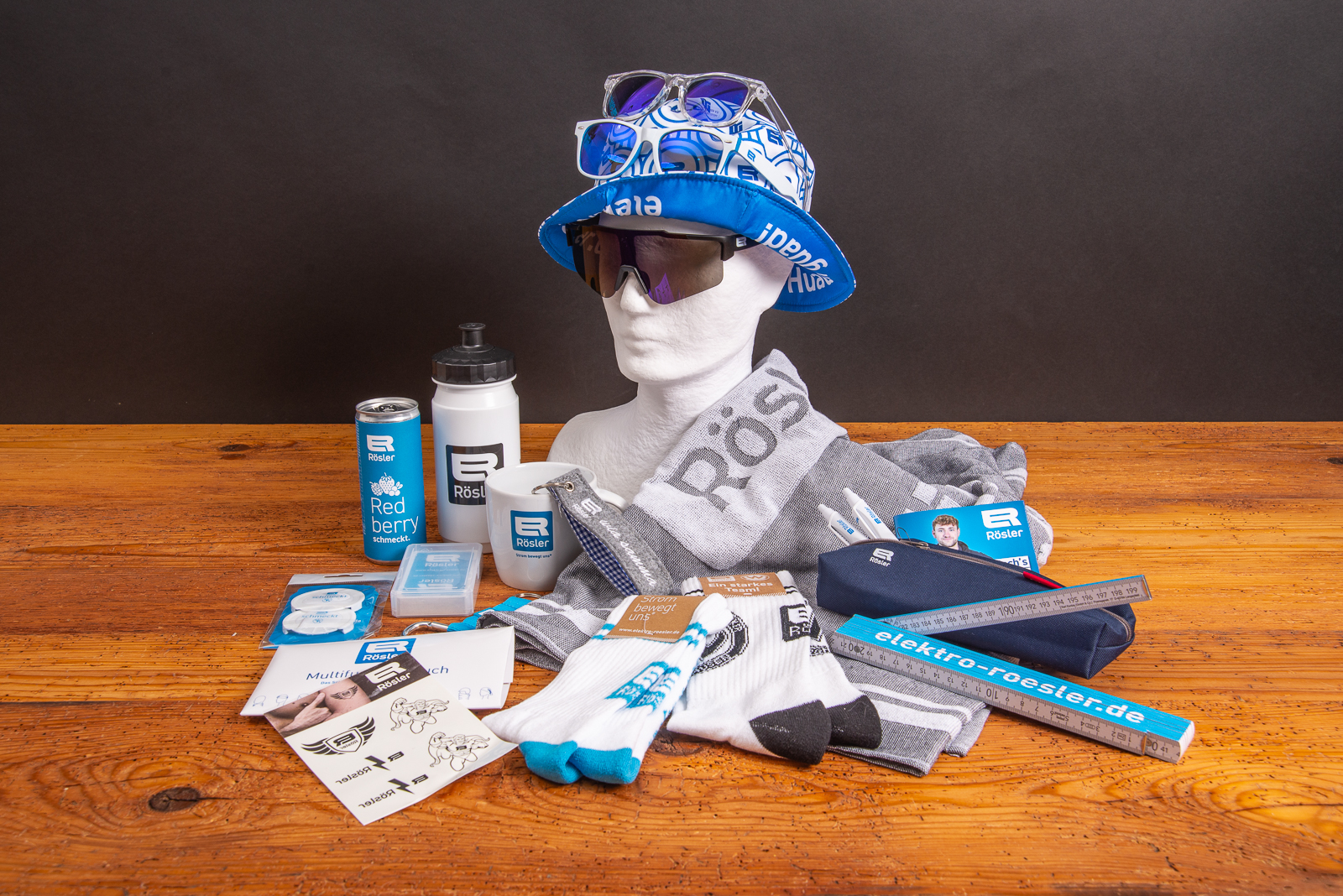 Merchandising-Set mit R&ouml;sler Werbeartikeln: bedrucktes Handtuch, Trinkflasche, Tasse, Socken, Sonnenbrillen, Bucket Hat, Lineal, Schreibwaren, Energy-Drink-Dose und weitere Sportfanartikel mit Logo auf Holzfl&auml;che arrangiert.