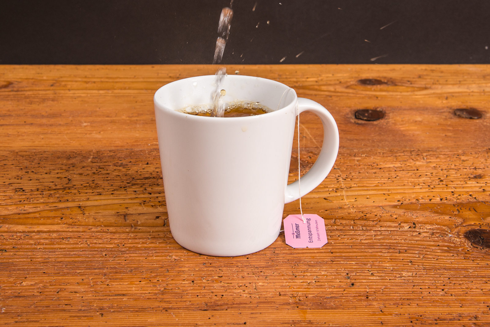 Wei&szlig;er SND Porzellanbecher mit Teebeutel aufgegossen mit hei&szlig;em Wasser als idealer Werbeartikel mit Logo bedruckt auf Holztisch.
