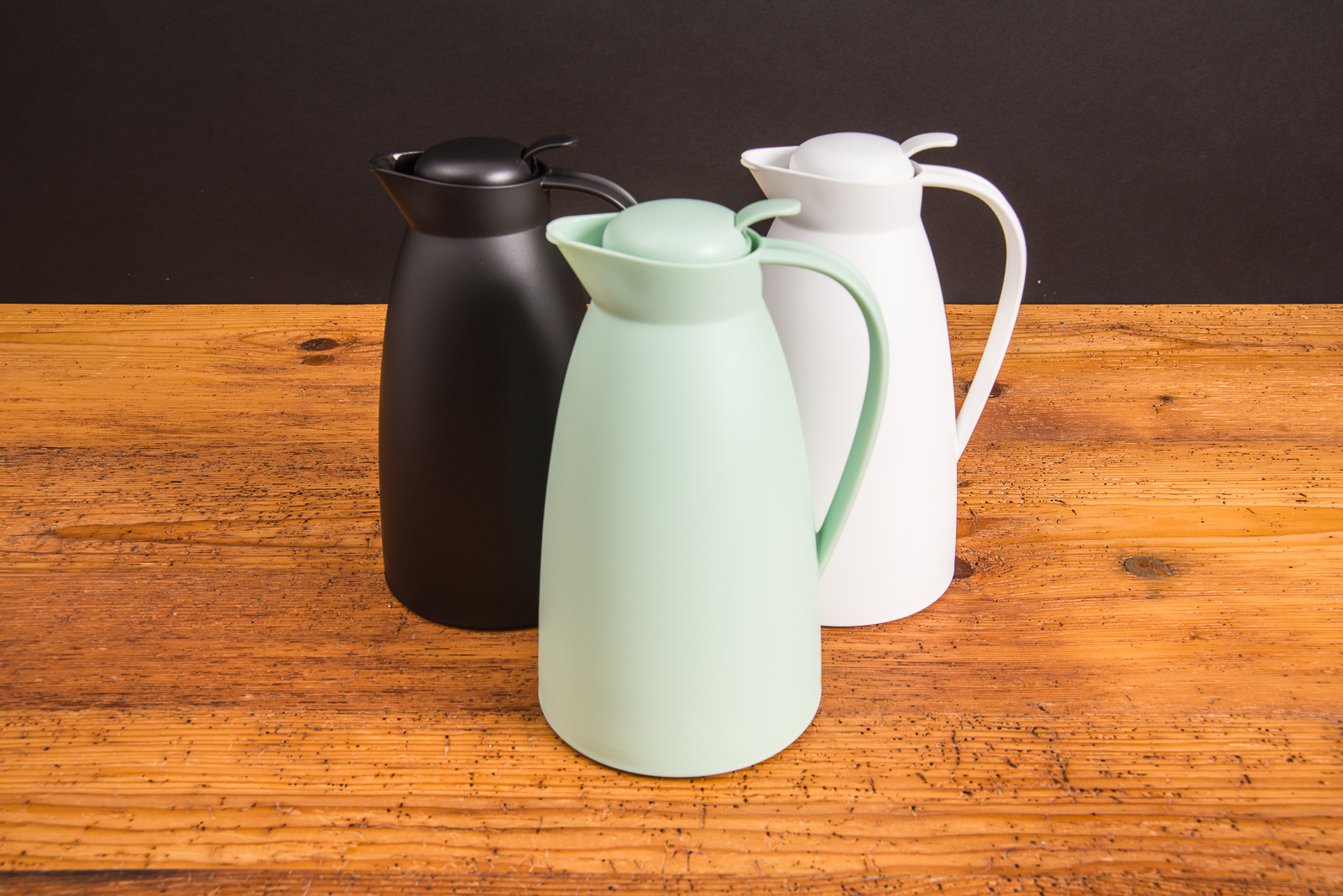 Drei alfi Isolierkannen ECO in Schwarz, Wei&szlig; und Mintgr&uuml;n auf Holztisch &ndash; stilvolle Thermoskannen als nachhaltige Werbeartikel mit Logo.