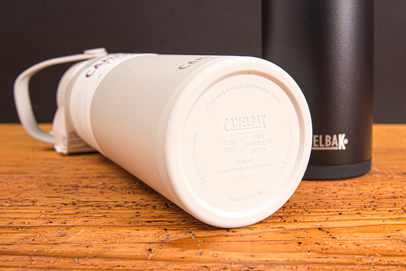 CamelBak Thrive VSS Trinkflaschen in Wei&szlig; und Schwarz &ndash; Detailansicht der Flaschenunterseite mit integriertem Protective Bumper f&uuml;r leisen und sicheren Halt auf nahezu jedem Untergrund  aus Edelstahl, hochwertige Thermoflaschen als nachhaltige Werbegeschenke mit Logo auf Holztischplatte.