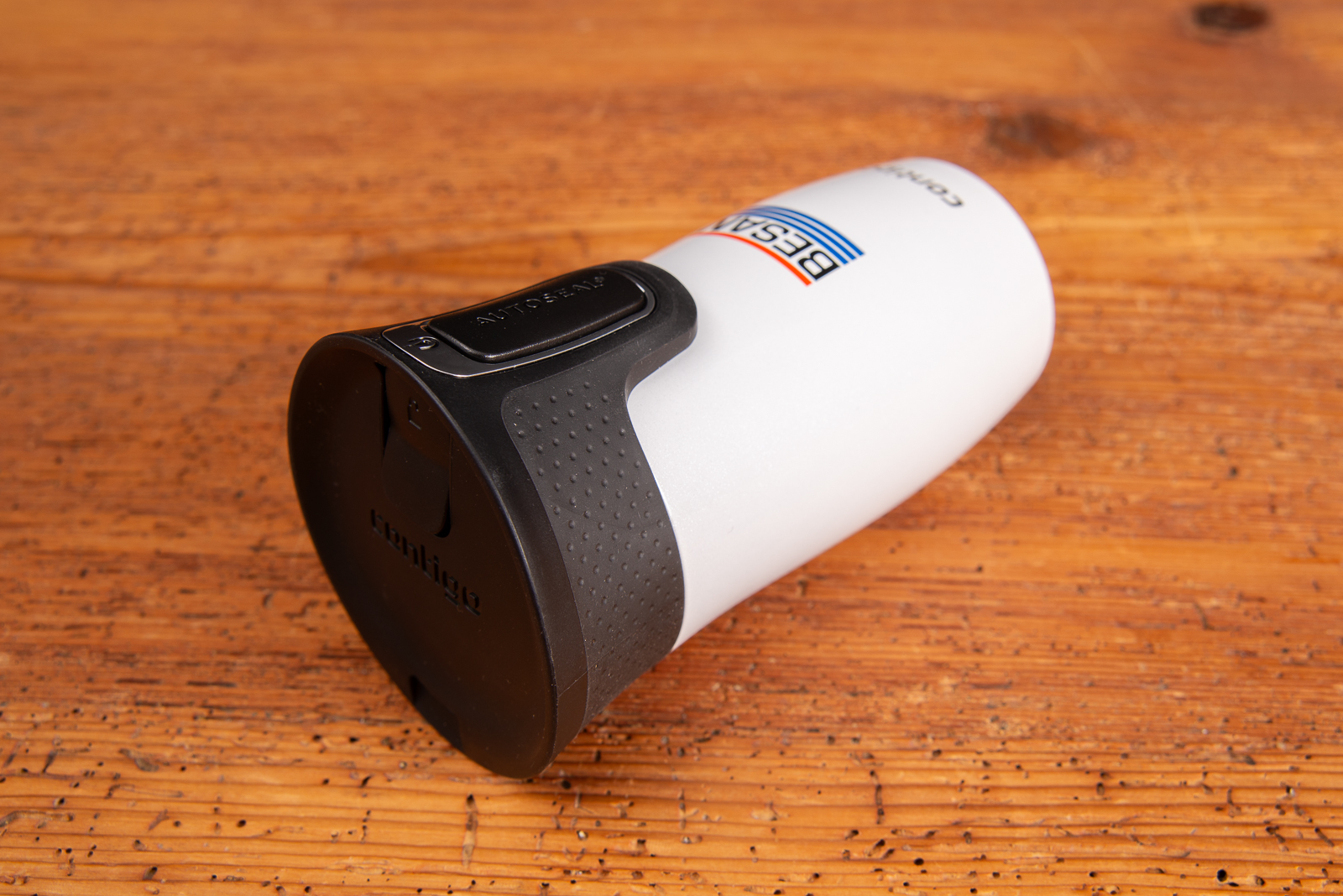 Contigo Thermobecher mit Besan Logodruck in Wei&szlig;, liegend auf Holztisch ist ein doppelwandiger Isolierbecher mit AUTOSEAL-Verschluss und gummierter Griffzone.