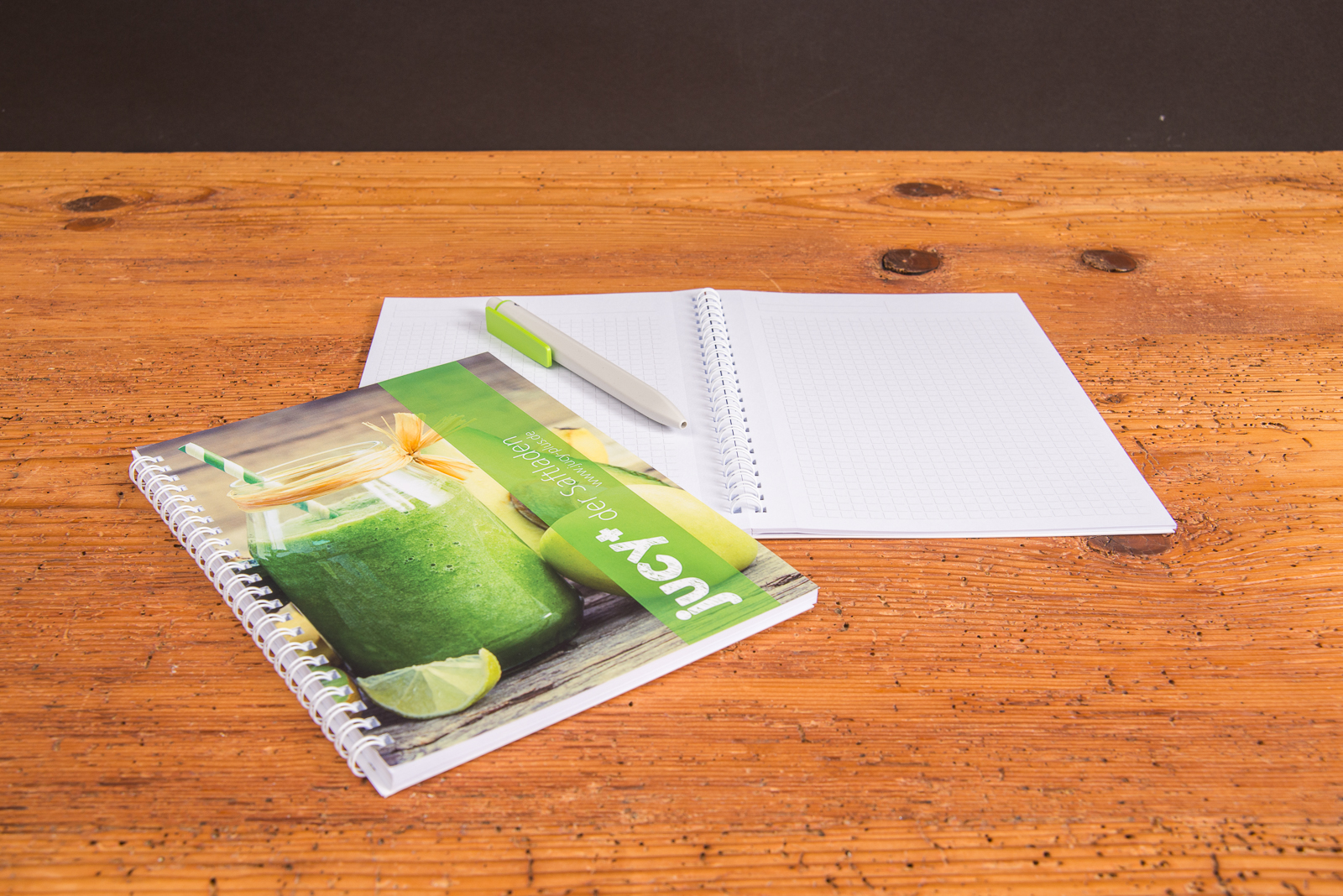 A5 Notizbuch mit Spiralbindung und individuell bedrucktem Cover im modernen Design, aufgeklappt auf Holztisch mit passendem Stift &ndash; ideales Meeting Book als g&uuml;nstiger Werbeartikel f&uuml;r Notizen, Seminare und Kundengespr&auml;che.