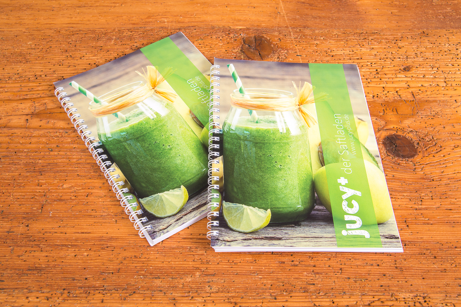 Zwei A5-Notizb&uuml;cher mit Spiralbindung und individuell bedrucktem Titelblatt im frischen Design mit gr&uuml;nem Smoothie-Motiv, abgebildet auf Holztisch &ndash; ideale, personalisierbare Werbeartikel f&uuml;r Meetings, Schulungen und Events.