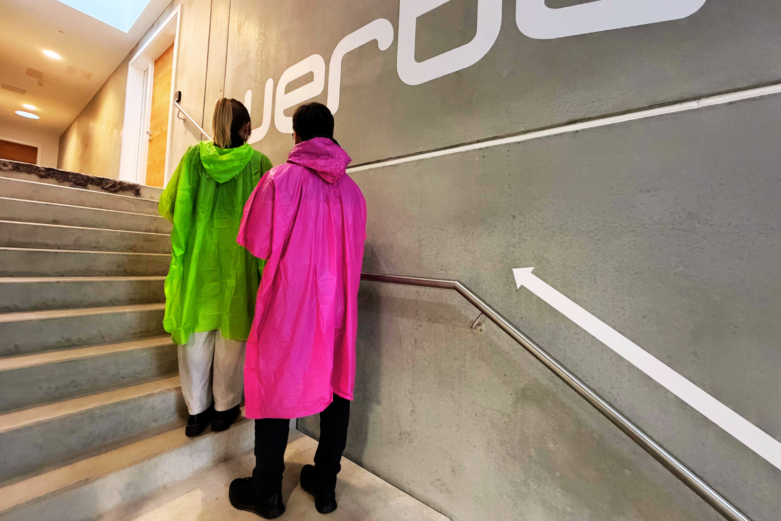 Zwei Personen tragen pinke und gr&uuml;ne Regenponchos aus biologisch abbaubarer Maisst&auml;rke und gehen eine Treppe hinauf &ndash; nachhaltige Regenponchos mit Logo als Werbeartikel f&uuml;r Festivals und Events von werbemax.