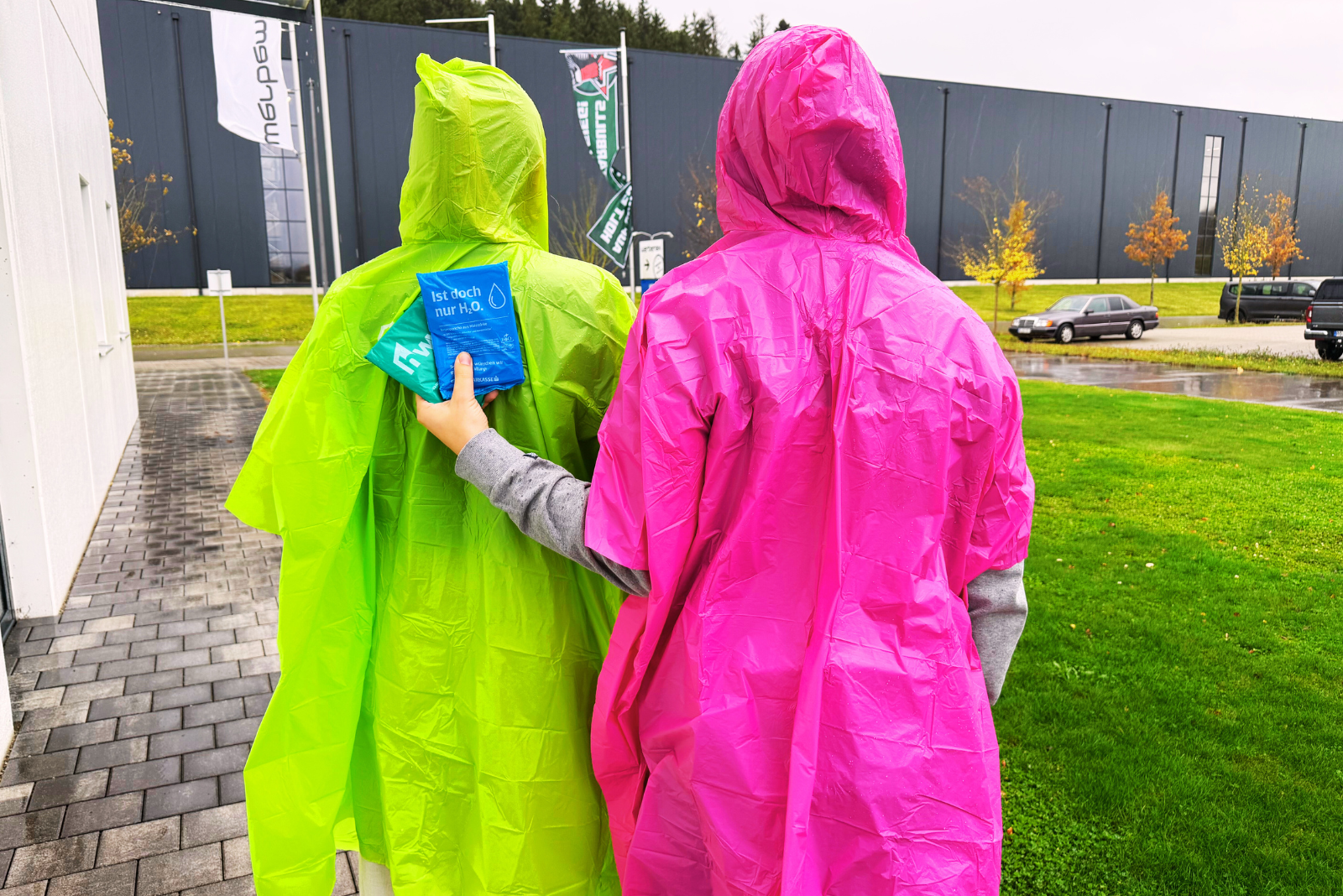 Zwei Personen mit gr&uuml;nen und pinken Regencapes aus biologisch abbaubarer Maisst&auml;rke gehen bei Regen &ndash; nachhaltige Regenponchos mit Logo als Werbeartikel f&uuml;r Festivals, Events und Tourismus von werbemax.