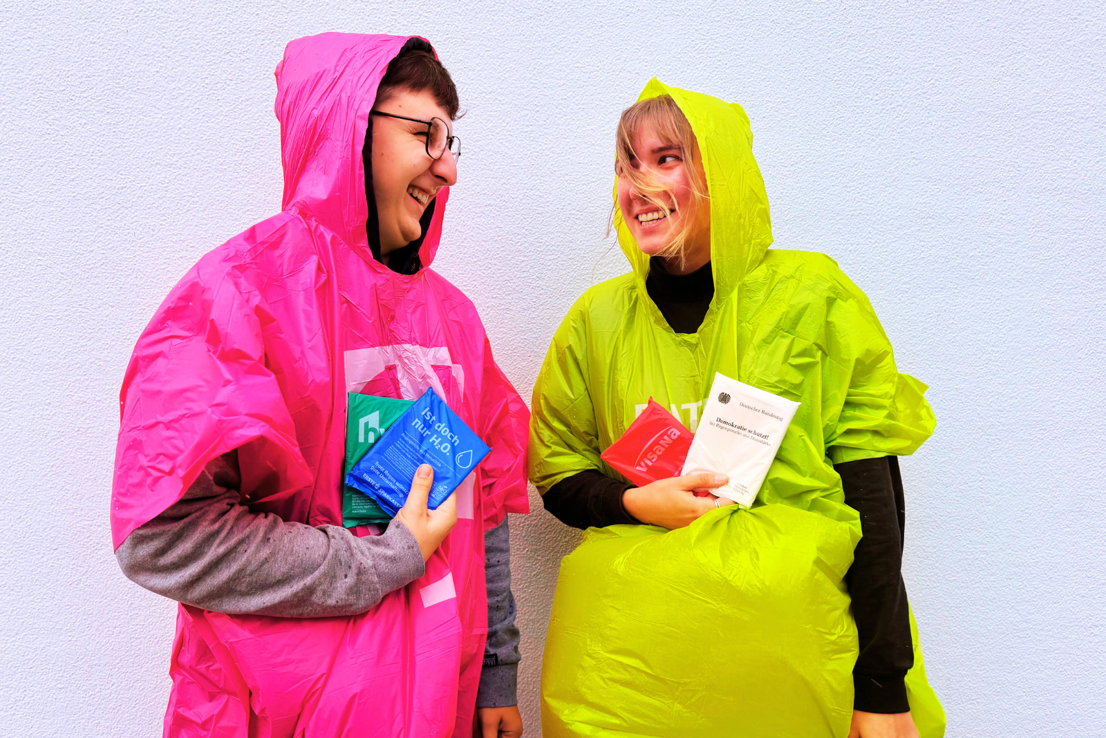 Zwei Personen tragen farbige Regenponchos aus biologisch abbaubarer Maisst&auml;rke mit Logo &ndash; nachhaltige Werbeartikel f&uuml;r Events, Festivals und Tourismusaktionen von werbemax.