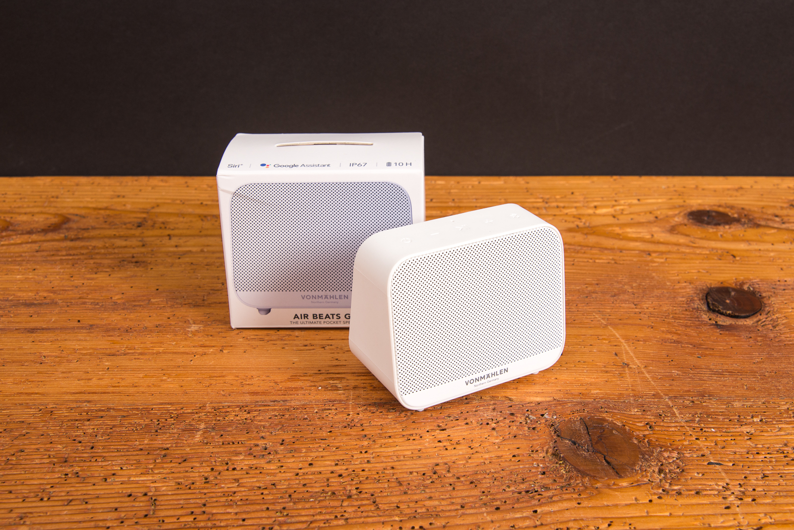 Bluetooth Box Air Beats Go in Wei&szlig; auf einem Holztisch stehend mit der Fachhandelsverpackung im Hintergrund. Der wei&szlig;e Karton mit der Produktabbildung dr&uuml;ckt nochmal Wertigkeit bei den Beschenkten aus. 