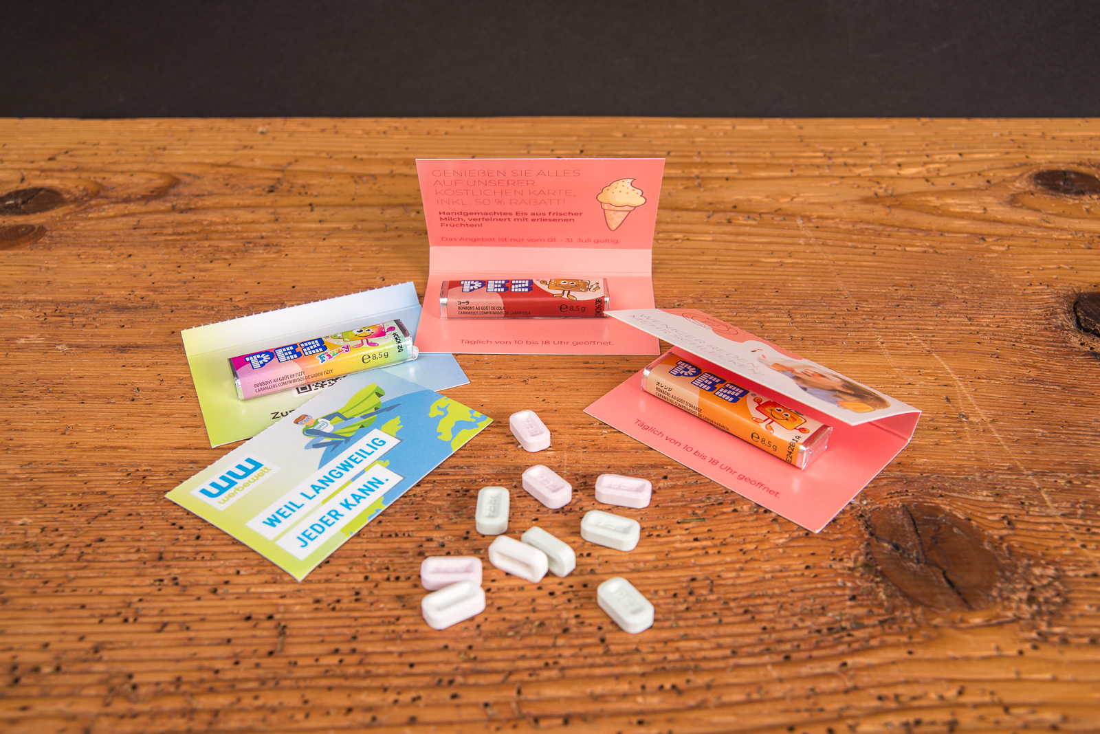 Verschiedene bedruckte PEZ-Werbekarten mit PEZ-Sticks als kreatives Give-away, pr&auml;sentiert mit Bonbons auf Holztisch.