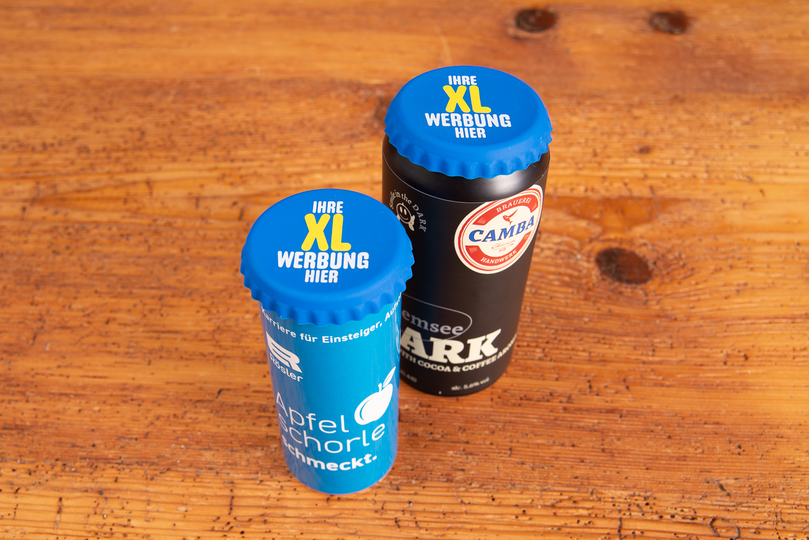 2 blaue, bedruckte XL Korki Can Silikon-Dosendeckel auf Getr&auml;nkedosen als praktisches Merch f&uuml;r Festivals, Events und Messen auf Holztisch stehend