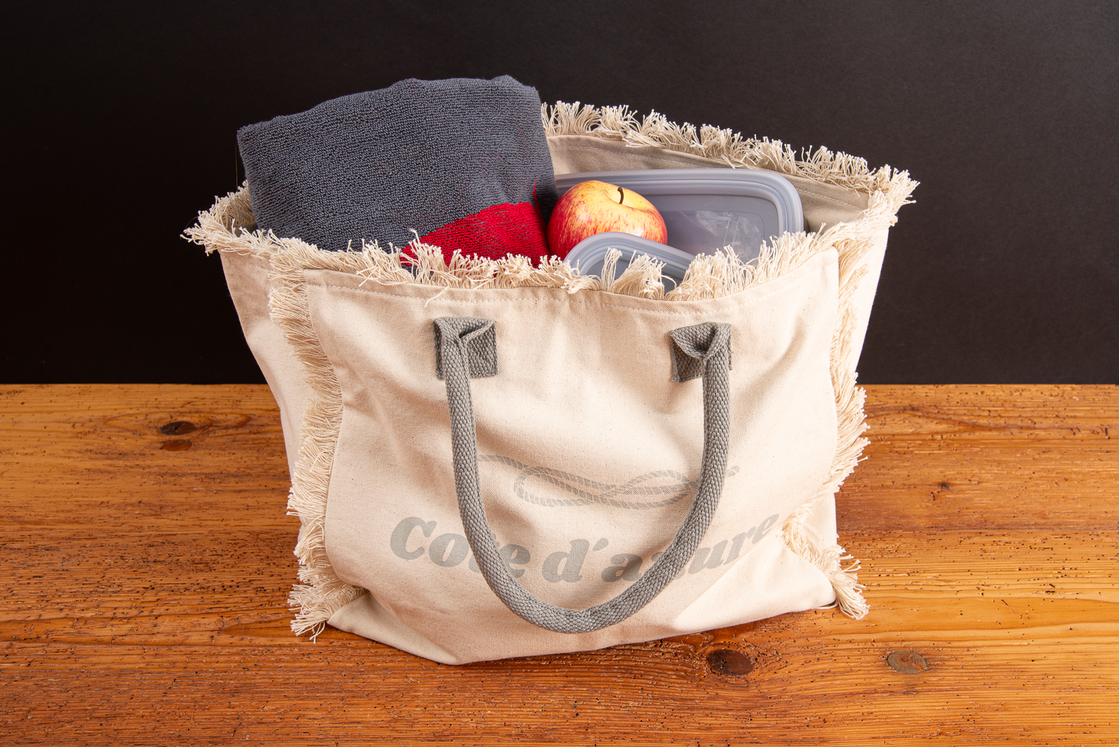 Strandtasche Lola als Werbeartikel &ndash; bedruckte Fairtrade-Badetasche mit grauen Griffen, gef&uuml;llt mit Strandutensilien.