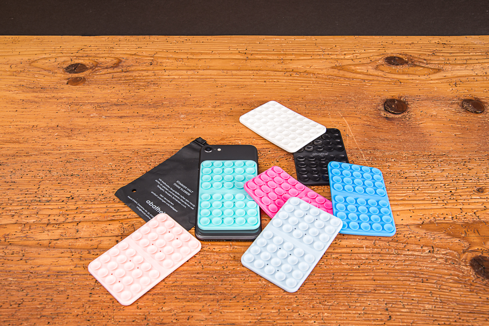 Produkt&uuml;bersicht der MagSquid Saugnapf-Halterung aus Silikon in Pastellrosa, T&uuml;rkis, Wei&szlig;, Schwarz, Pink und Blau neben einer schwarzen Polybeutel-Verpackung.
