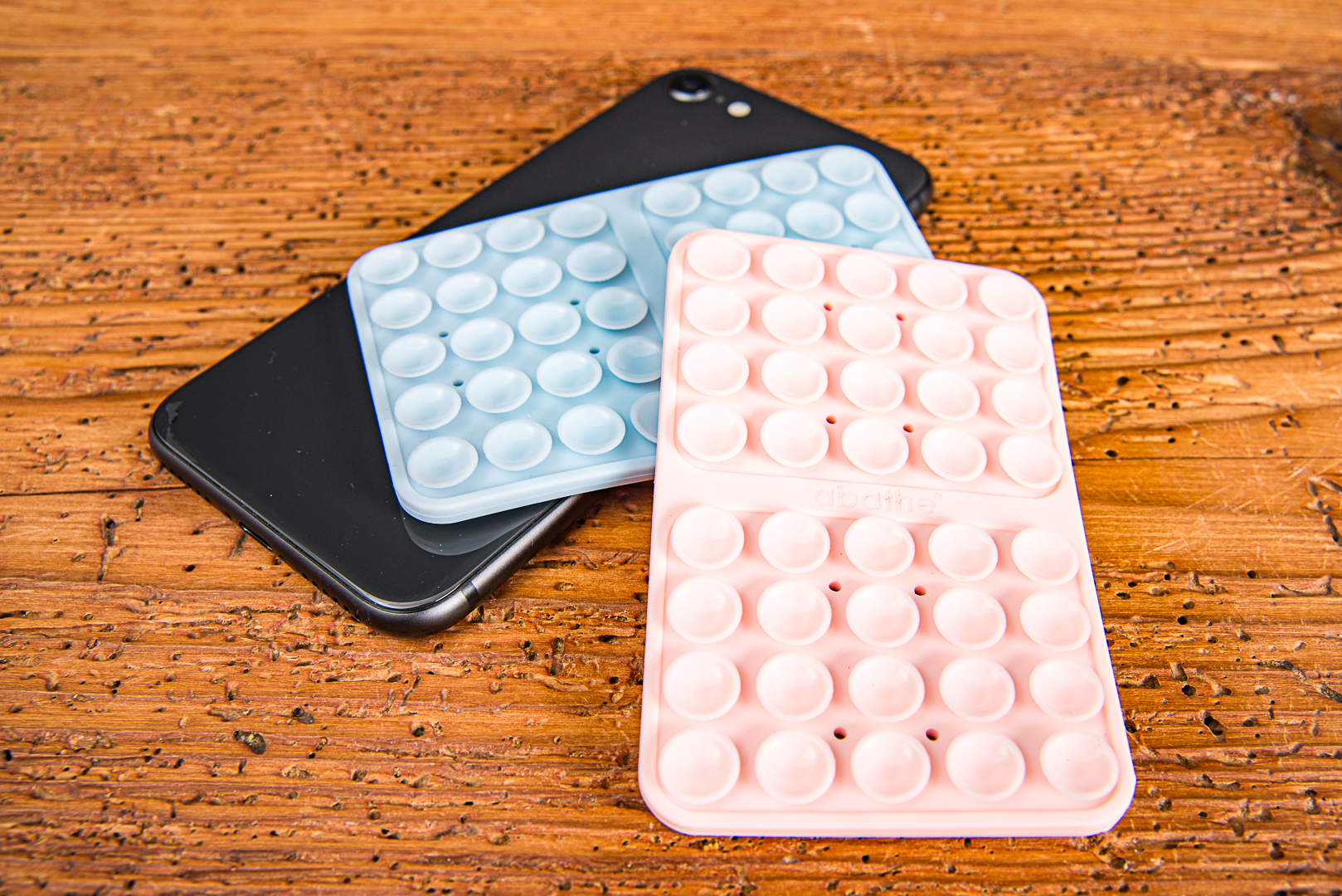 MagSquid Two Sides Saugnapf-Handyhalterungen in Pastellblau und Pastellrosa auf einem schwarzen Smartphone auf einem Holztisch.