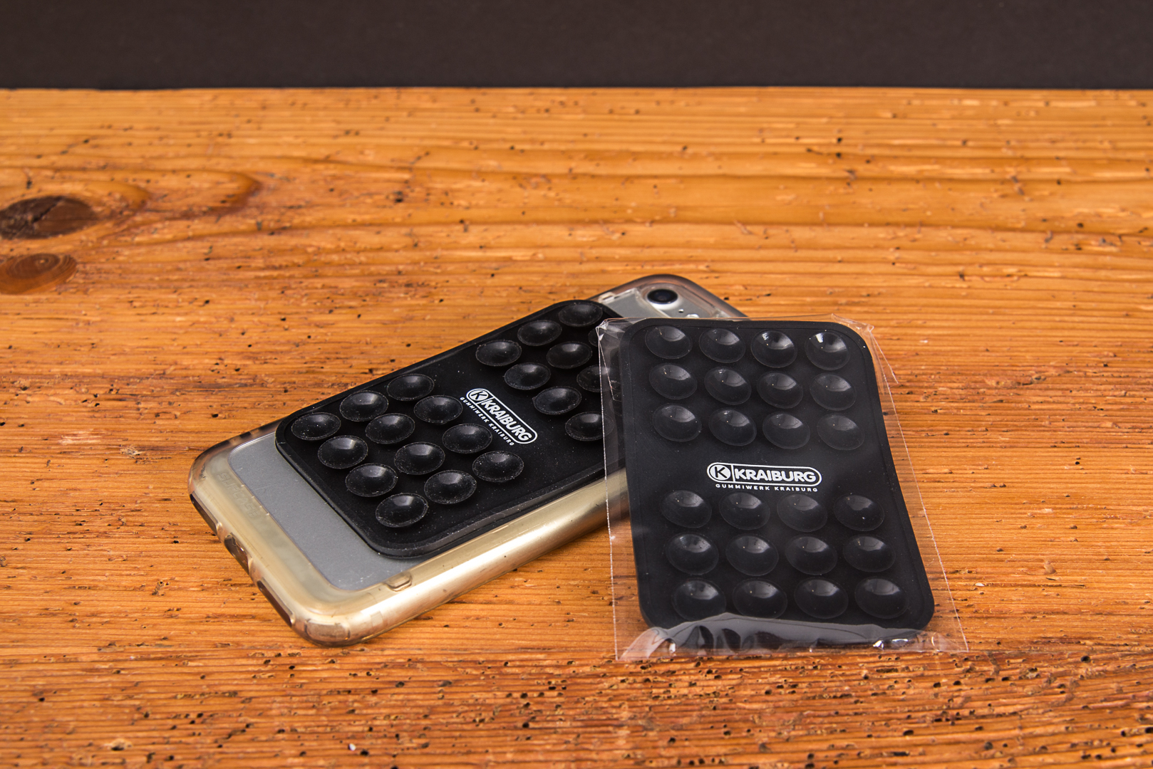 MagSquid Premium Silikon-Saugnapf-Pad in Schwarz mit Logo-Lasergravur auf einem Handy. Werbemittel einzeln verpackt in OPP-Folie auf einem Holztisch.