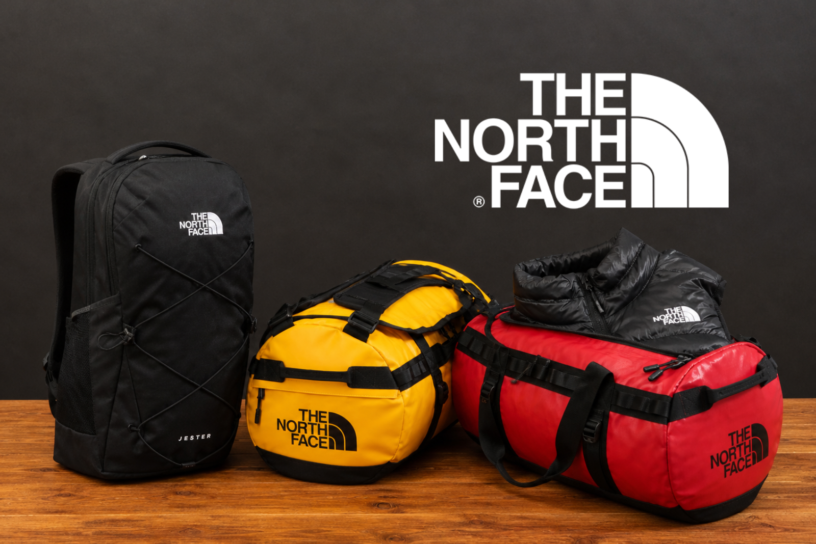 The North Face Werbeartikel mit Logo: Rucksack Jester, Base Camp Duffel Bags und Terra Peak Weste als hochwertige Markenbotschafter auf Holztisch inszeniert.