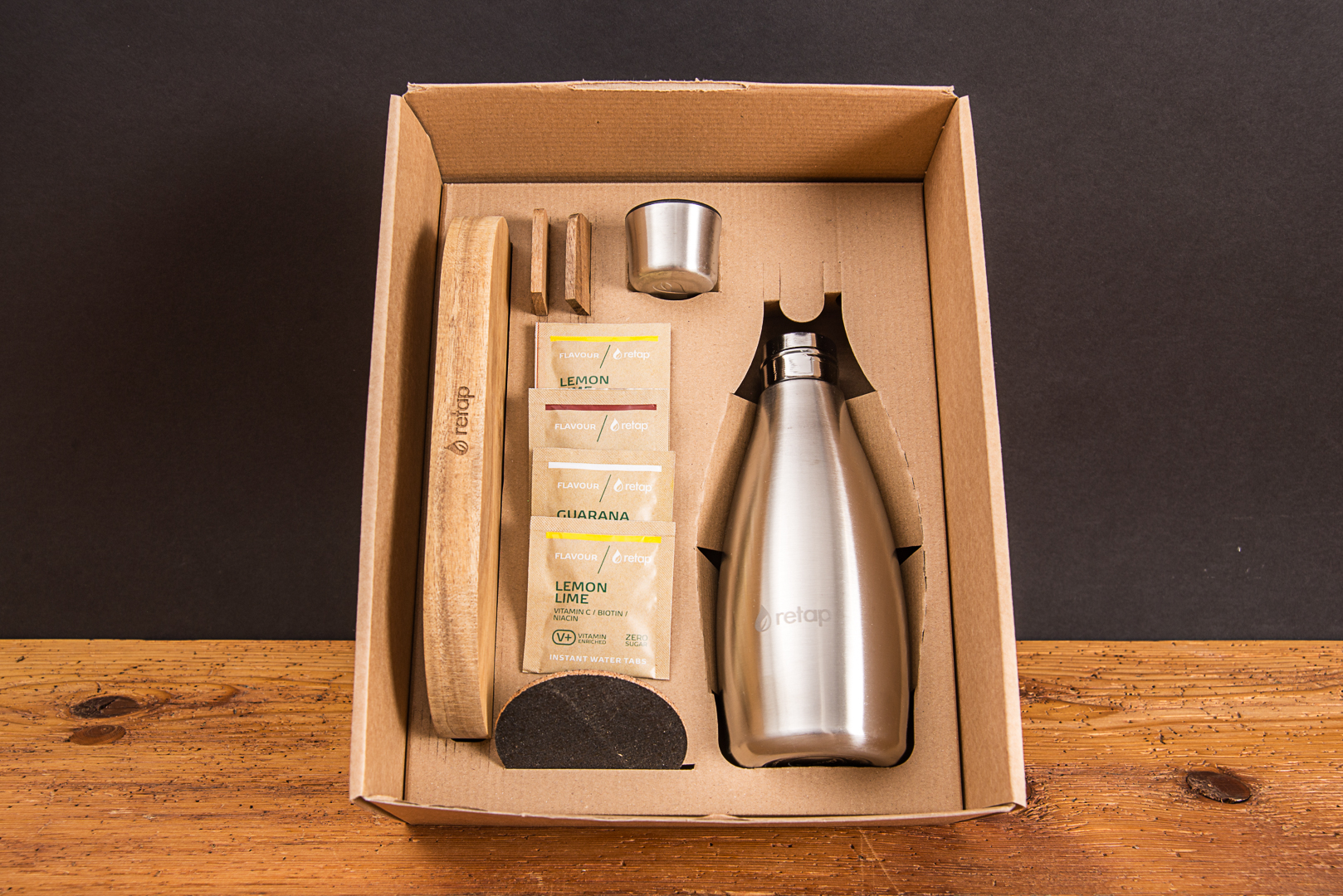 Ge&ouml;ffnete Geschenkbox f&uuml;r neue Mitarbeiter mit retap Wooden Tray aus Eichenholz, einer Edelstahl-Trinkflasche und Flavours als hochwertiges Onboarding-Pr&auml;sent von werbemax.