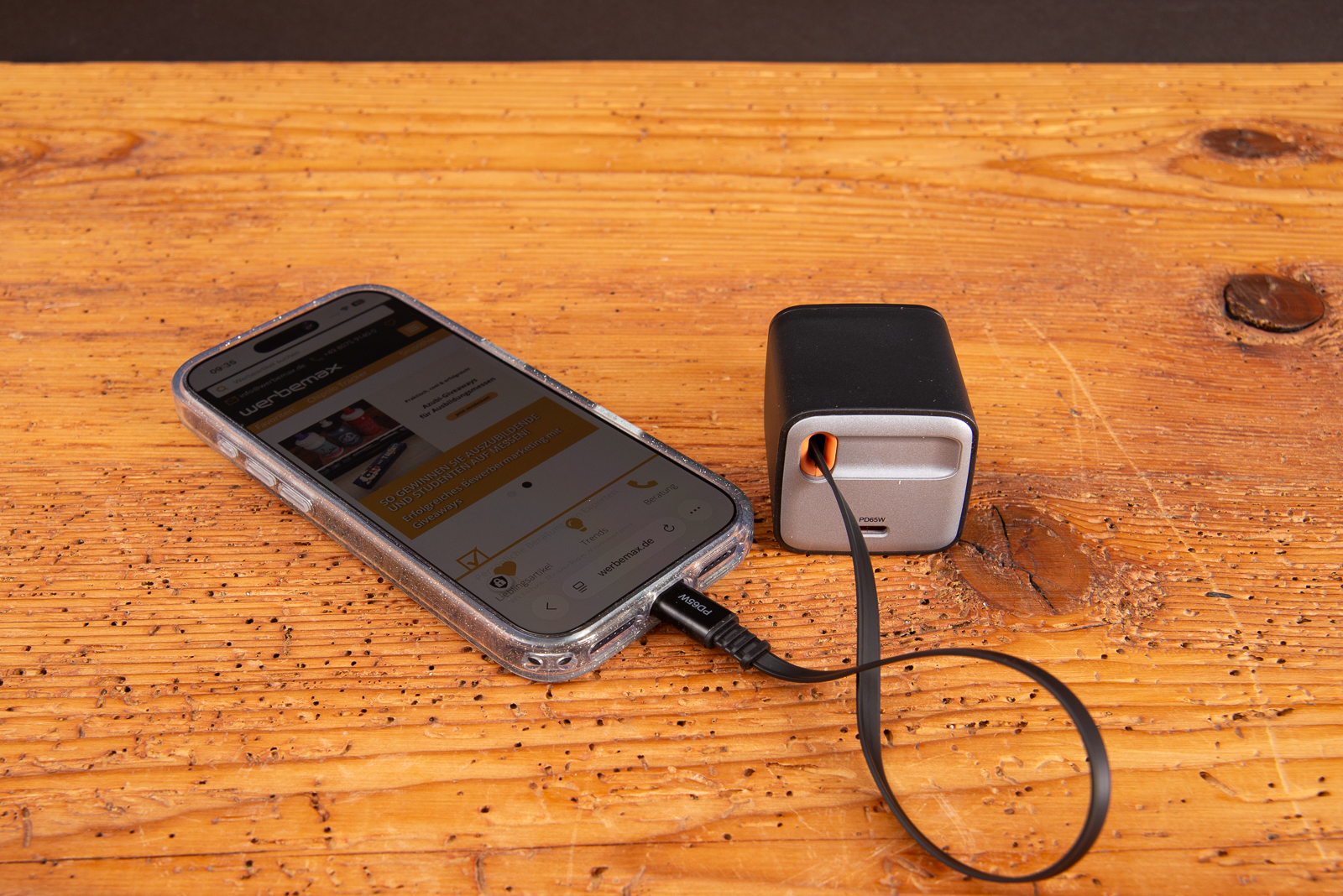 werbemax SUPPLY 65 C GaN Ladeger&auml;t 65W beim Laden eines Smartphones auf einem Holztisch &uuml;ber das integrierte USB-C Ausziehkabel