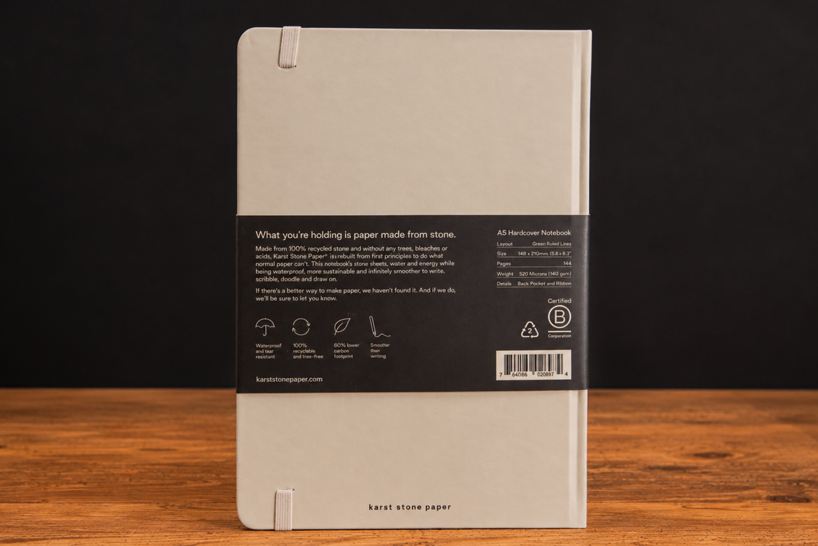 Nachhaltiges A5 Notizbuch aus Stone Paper auf Holztisch vor schwarzem Hintergrund &ndash; umweltfreundliches Hardcover-Notebook mit Steinpapier und Informationsbanderole.