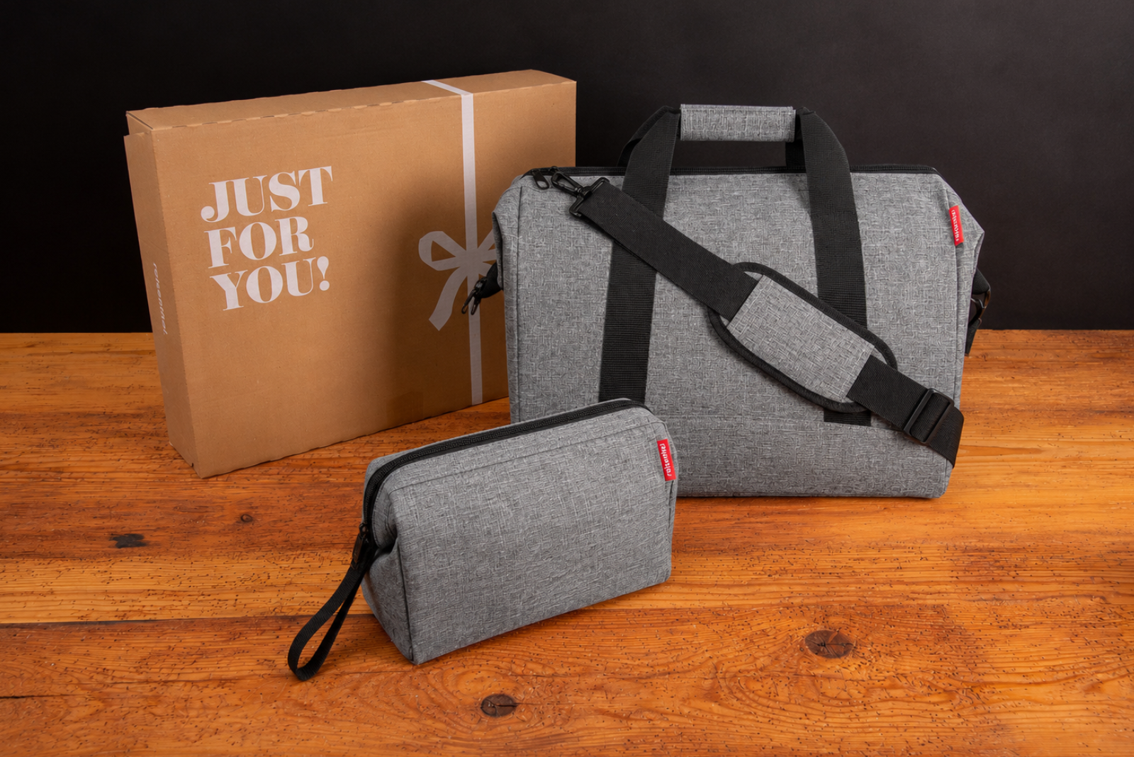 Graue Reisenthel Reisetasche mit Beautycase und Geschenkkarton als hochwertiges Werbegeschenk auf rustikalem Holztisch.
