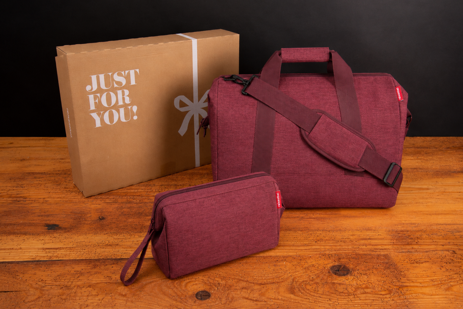 Bordeauxrote Reisenthel Reisetasche mit Kosmetiktasche und Geschenkbox auf Holztisch vor dunklem Hintergrund.