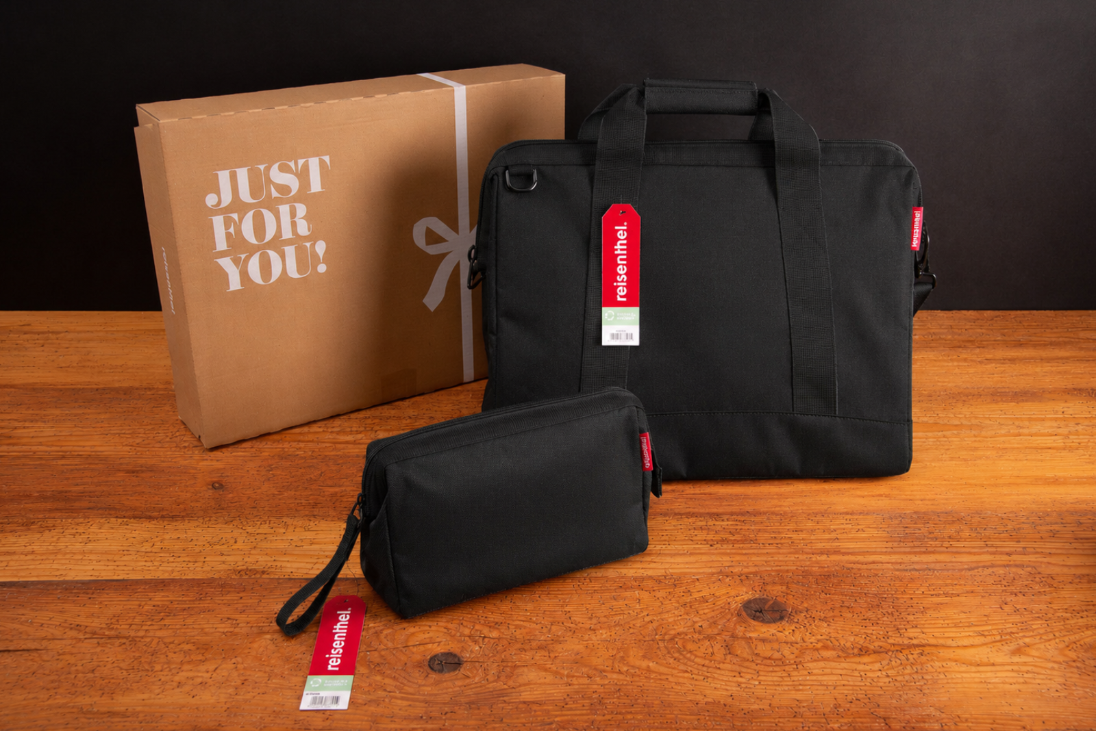 Schwarze Reisenthel Reisetasche mit passendem Beautycase als Geschenkset auf Holztisch vor schwarzem Hintergrund mit Geschenkkarton.