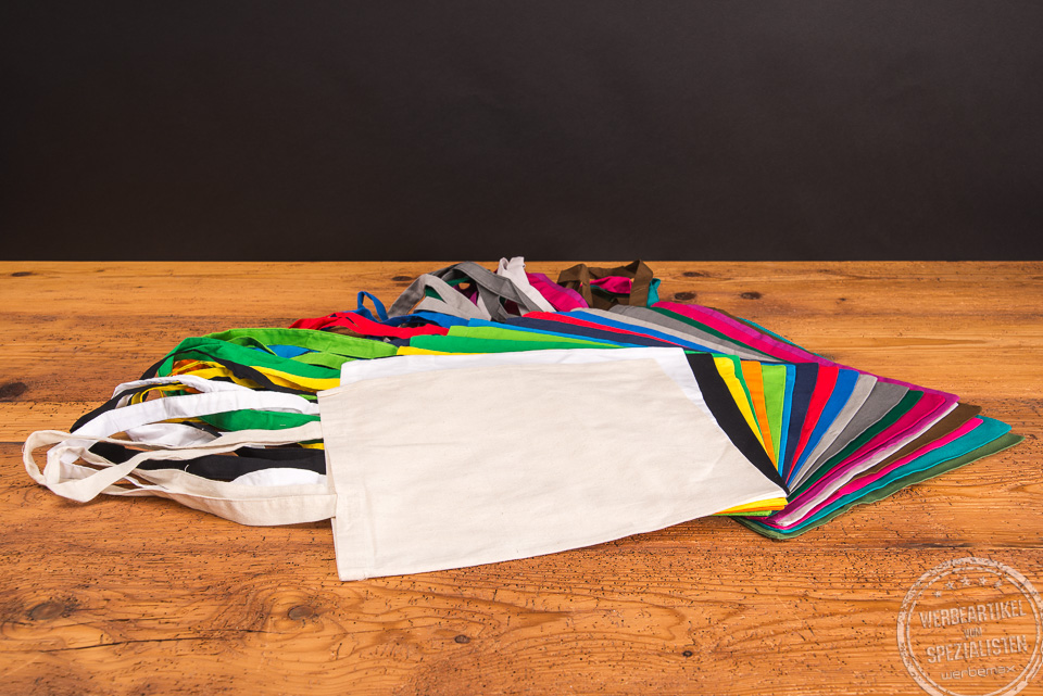 Messetasche in 22 Farben &uuml;bereinander aufgef&auml;chert liegend auf Holztisch als Give-away f&uuml;r Kunden