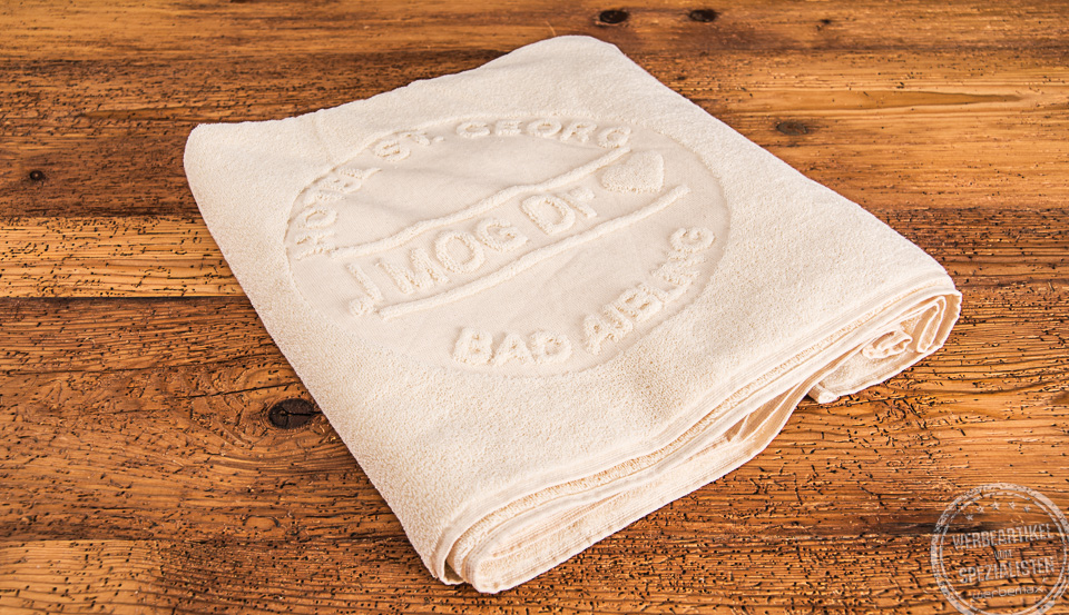 Beiges zusammengefaltetes UNDYED Handtuch mit Logo des Hotel Sankt Georg aus Bad Aibling als Werbegeschenk.