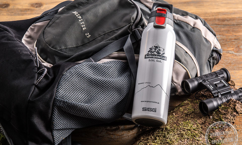 SIGG Trinkflasche Hot and Cold in der Farbe grau mit Berchtesgardener Land Aufdruck liegend auf einem Rucksack als Werbegeschenk.